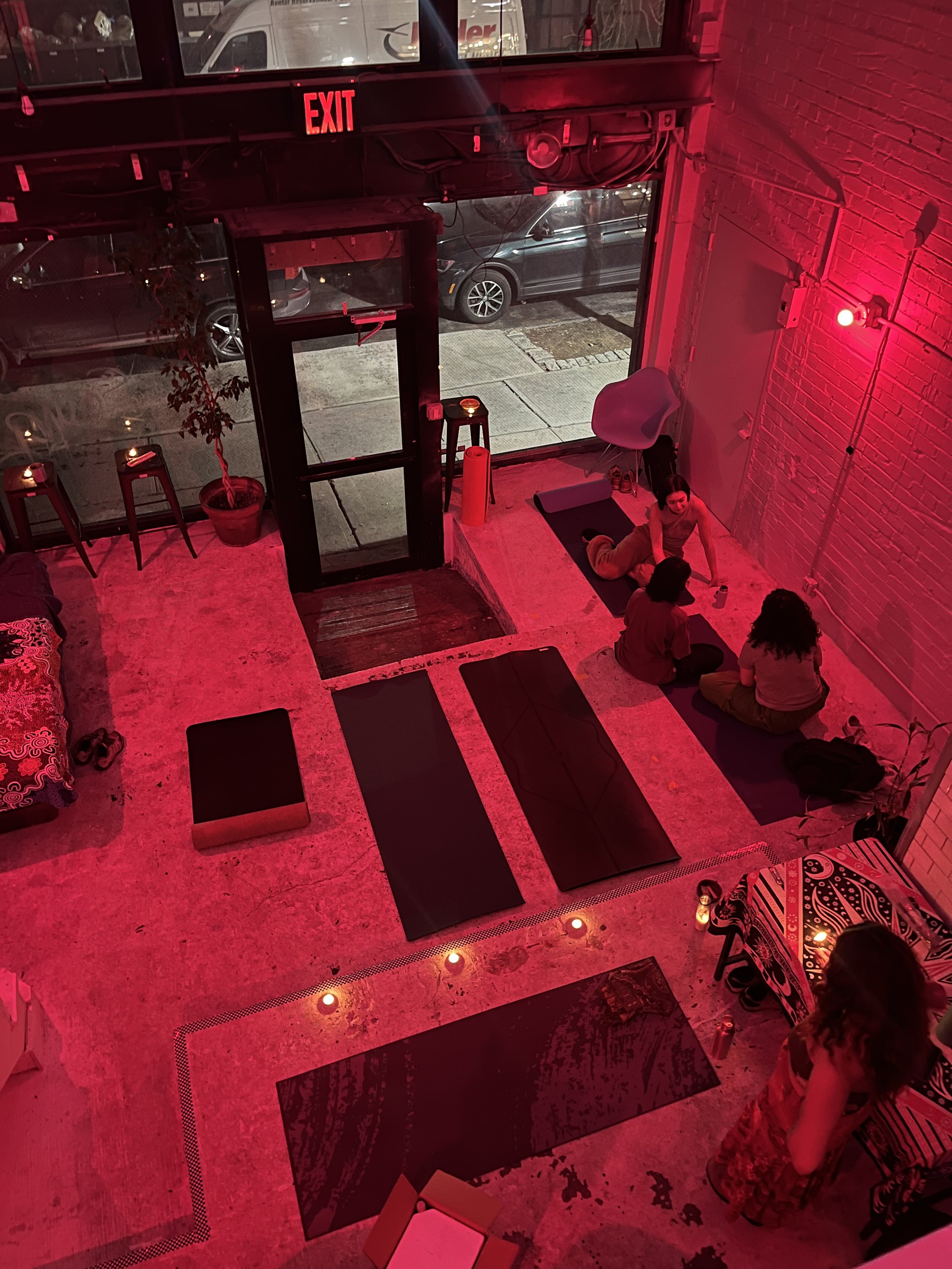 Indoor space with yoga mats on a concrete floor, low lighting, and red ambient lighting. Three people sit on two of the mats near the brick wall, and a woman stands nearby. Outside the large front window.