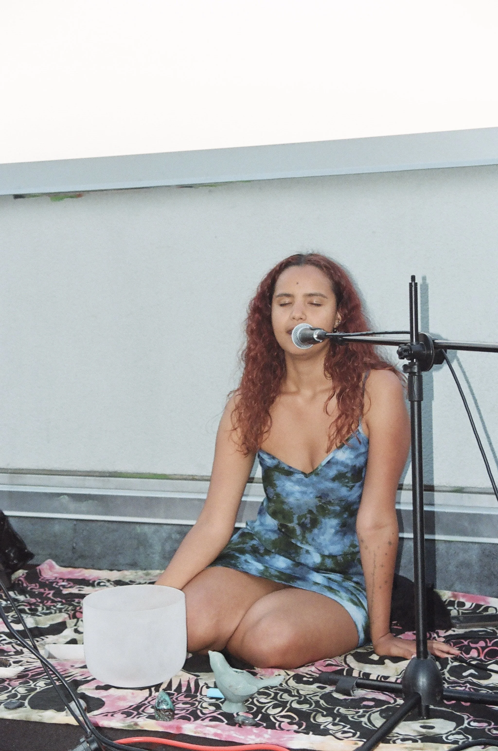 A young woman with curly red hair sitting cross-legged on a patterned blanket, singing into a microphone, with a white crystal singing bowl and small decorative items beside her.