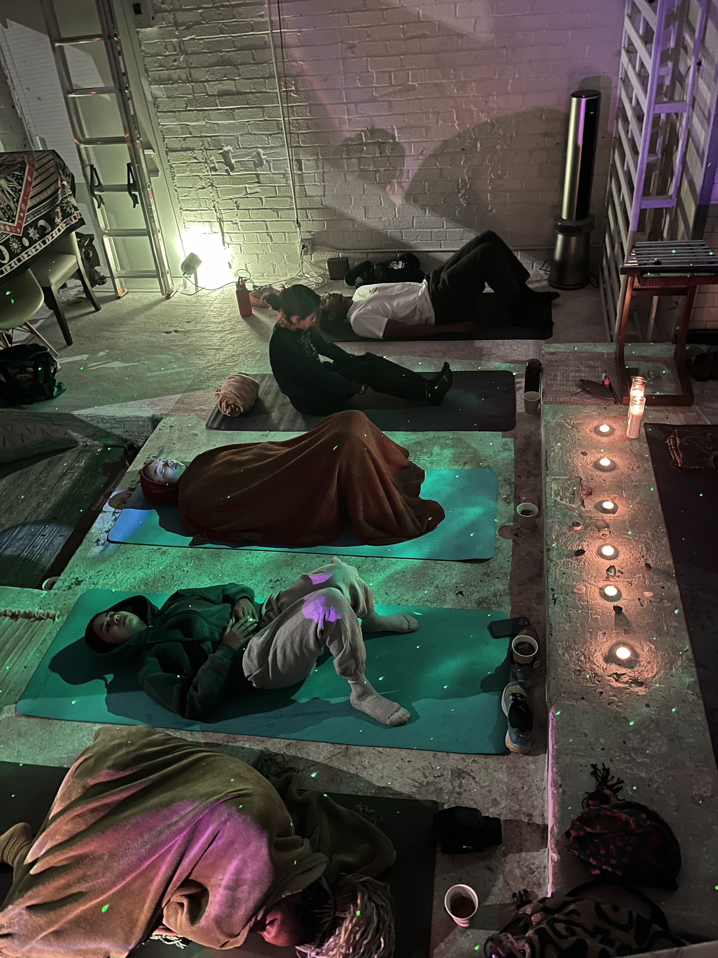 Group of people lying on yoga mats in dimly lit room, likely participating in a relaxation or meditation session, with candles and soft lighting creating a calming atmosphere.