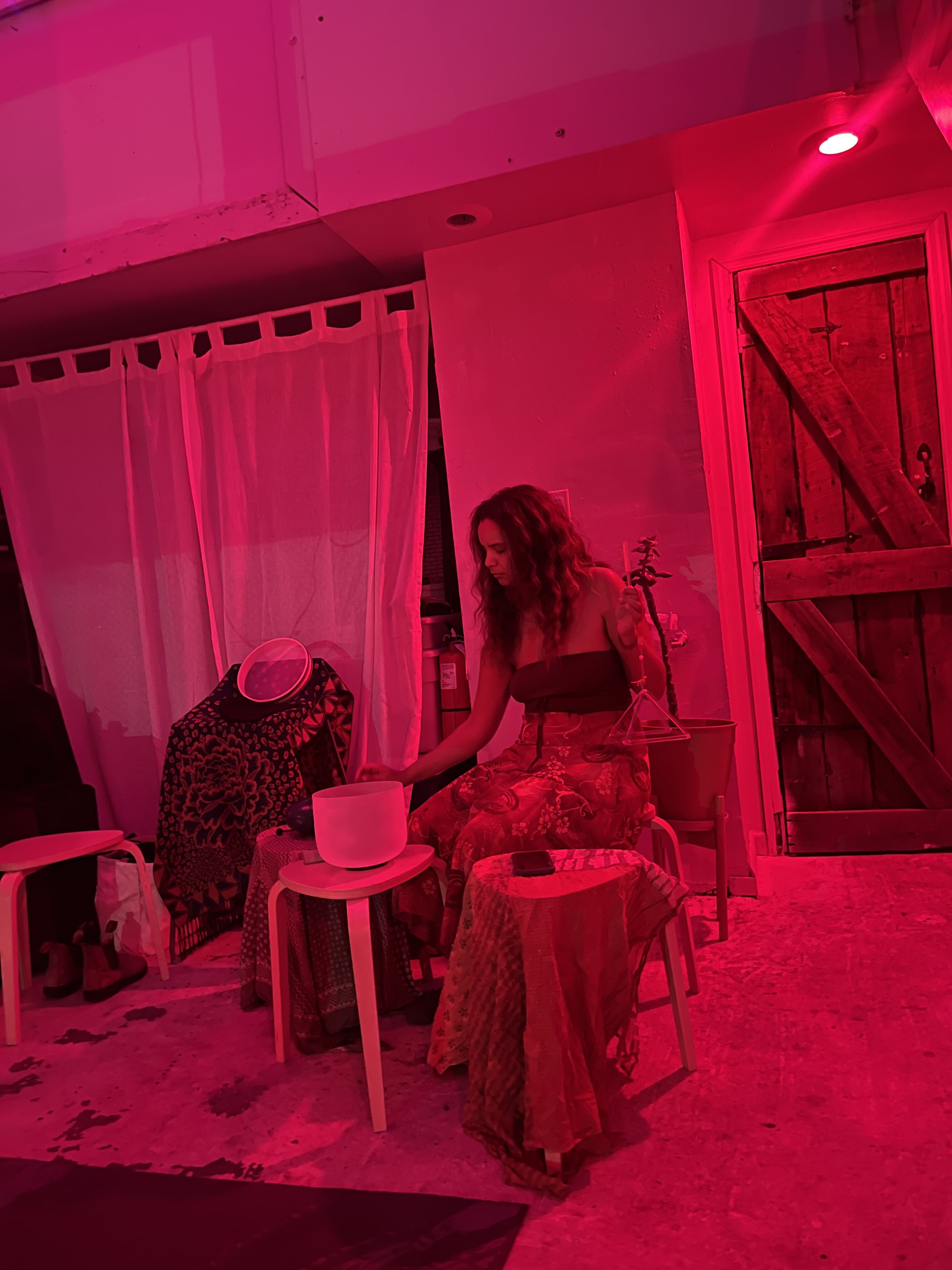 A woman with curly hair sits on a small chair in a room bathed in pink light, with a wooden door and pink curtains behind her. She appears to be playing a singing bowl or similar object.