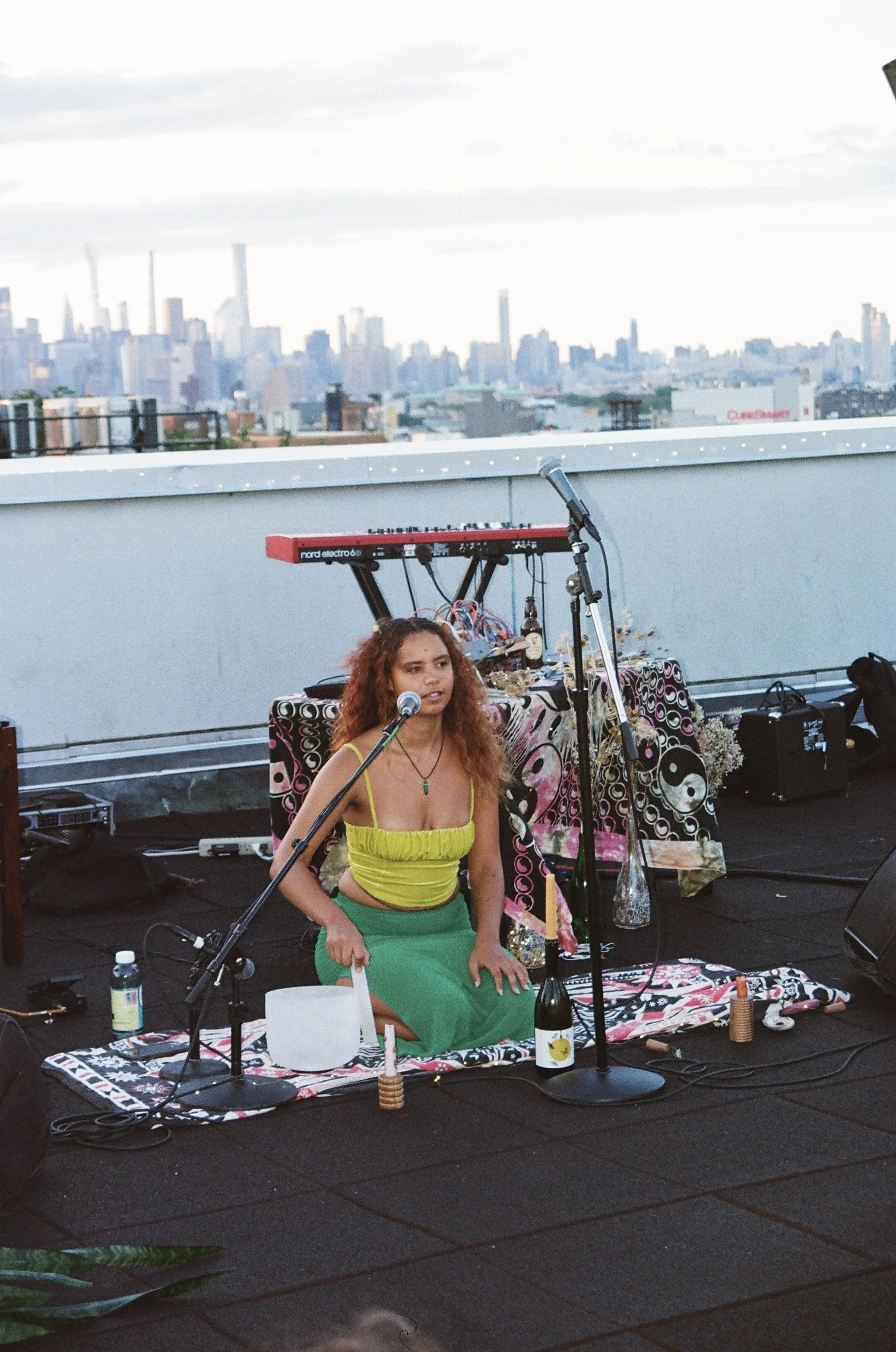 A woman with curly hair wearing a yellow top and green skirt sitting on a mat on a rooftop stage, surrounded by musical equipment, with a city skyline in the background during daytime.