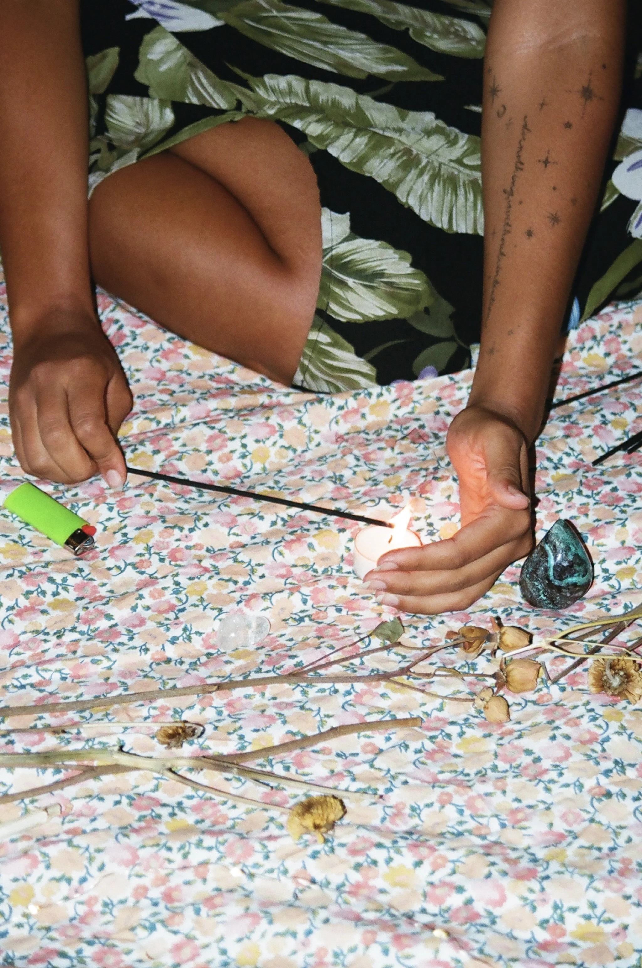 Person with tan skin and tattoos lighting a candle on a floral cloth with dried flowers and stones around.