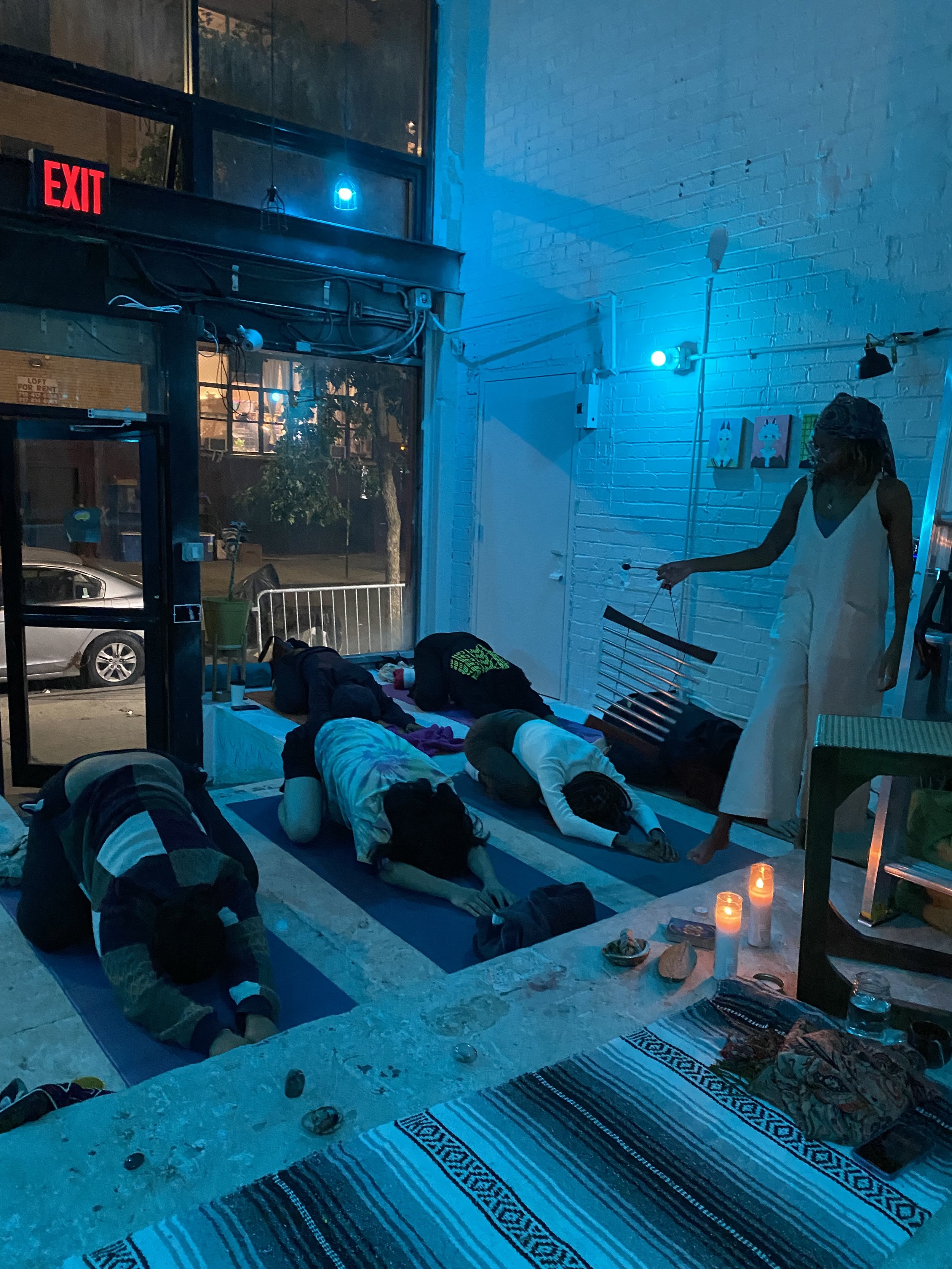 A group of people practicing yoga in a dimly lit, blue-toned indoor space with a white brick wall. A woman stands nearby guiding the session. Candles and small objects are on a table in the foreground.