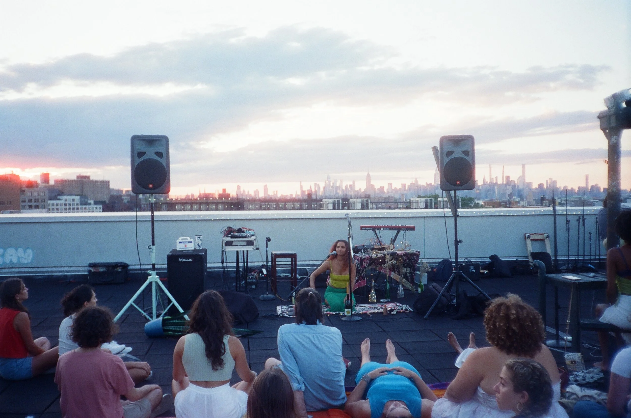 A rooftop gathering with a female performer sitting on a cushion, surrounded by an audience, with city skyline in the background during sunset.
