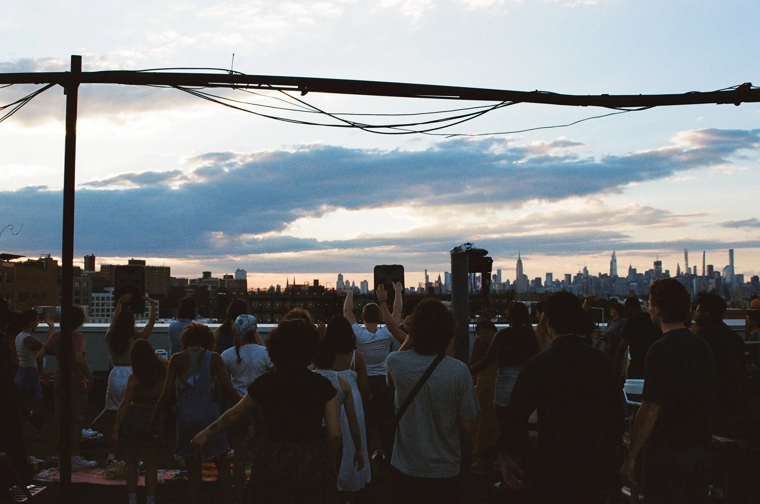 A group of people gathered on a rooftop at sunset with the New York City skyline in the background.