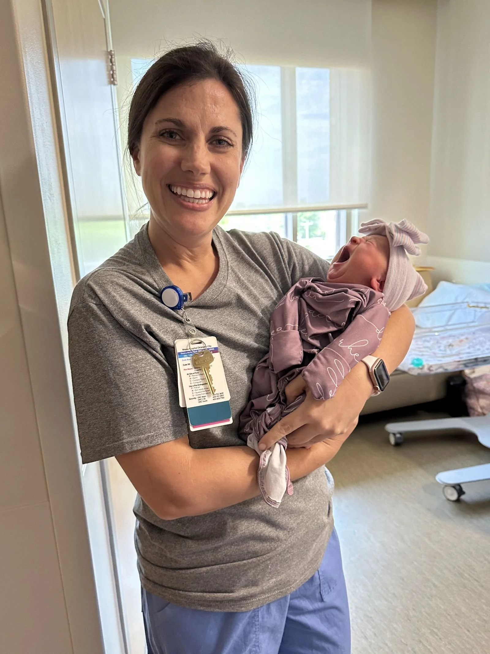 A woman in a gray shirt holding a newborn baby who is yawning in a hospital room.