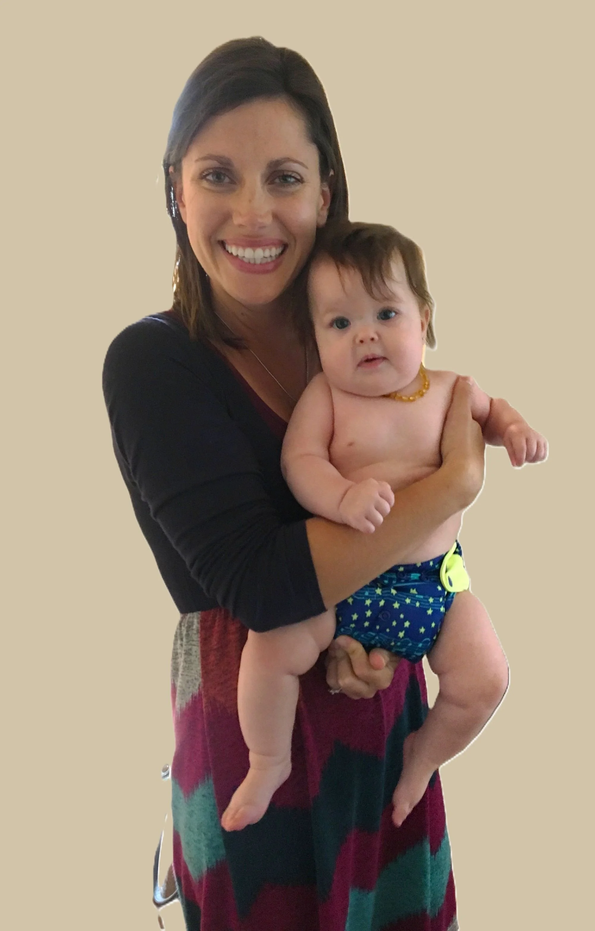 A woman with short brown hair smiling while holding a baby with light brown hair and blue eyes, who is shirtless and wearing a diaper and necklace, against a plain background.
