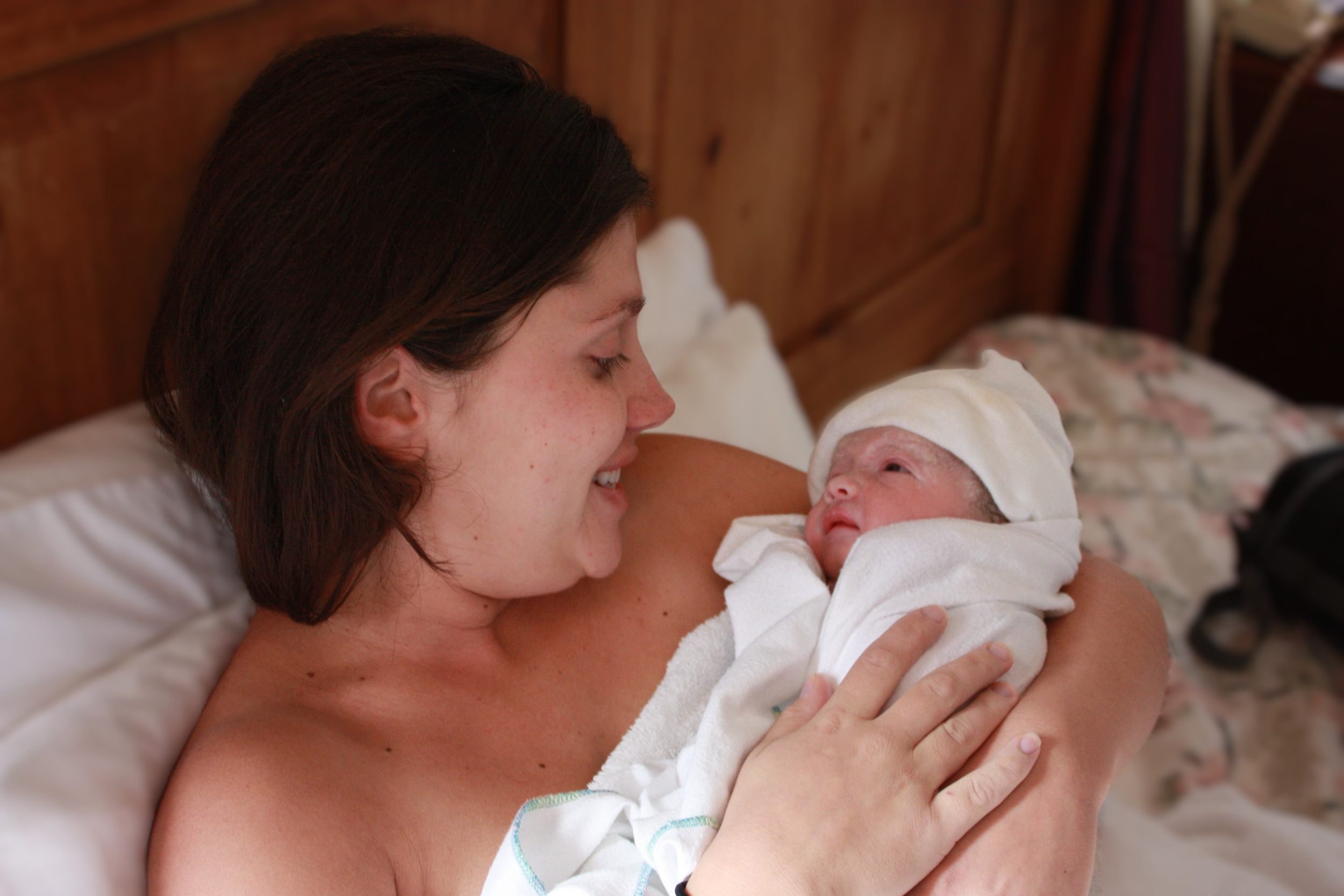 A woman holding a newborn baby wrapped in a white towel and wearing a white hat, looking at the baby with a smile.