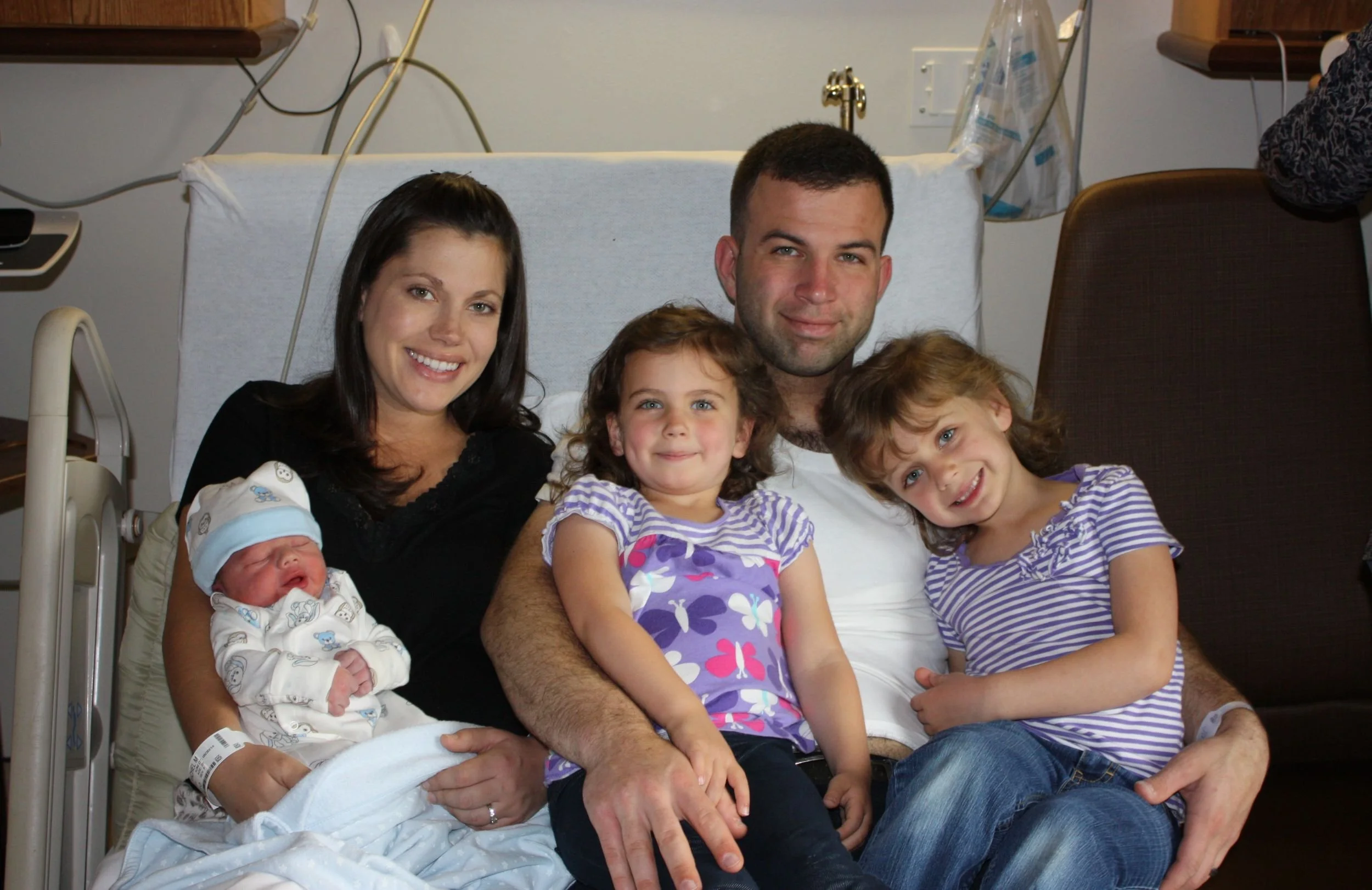 Hospital family photo showing a woman, man, two young girls, and a newborn baby in a hospital room.