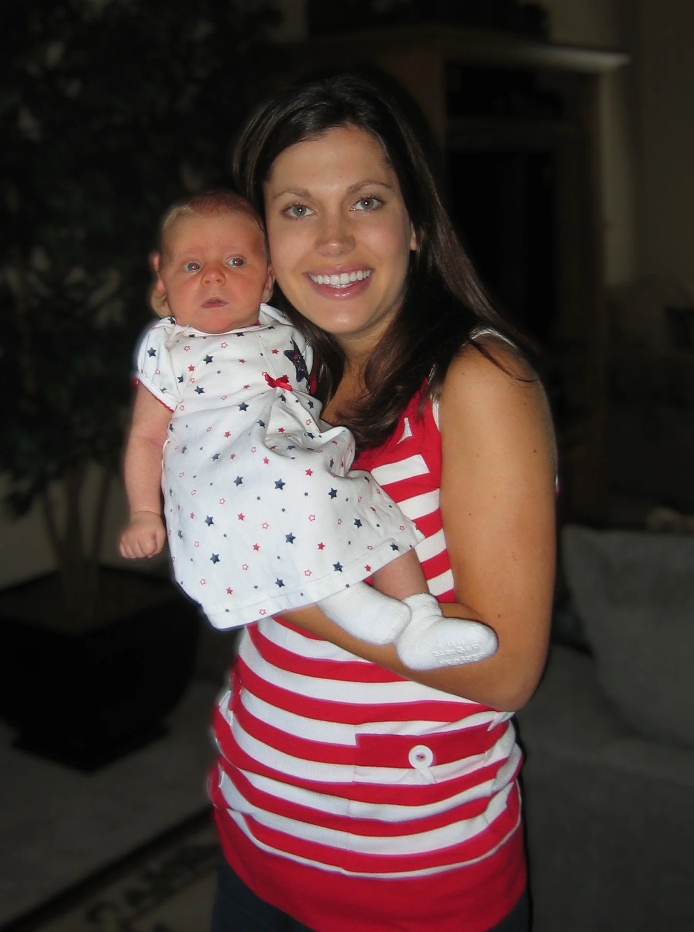 A woman holding a baby indoors, both wearing patriotic clothing with red, white, and blue colors and stars, smiling at the camera.