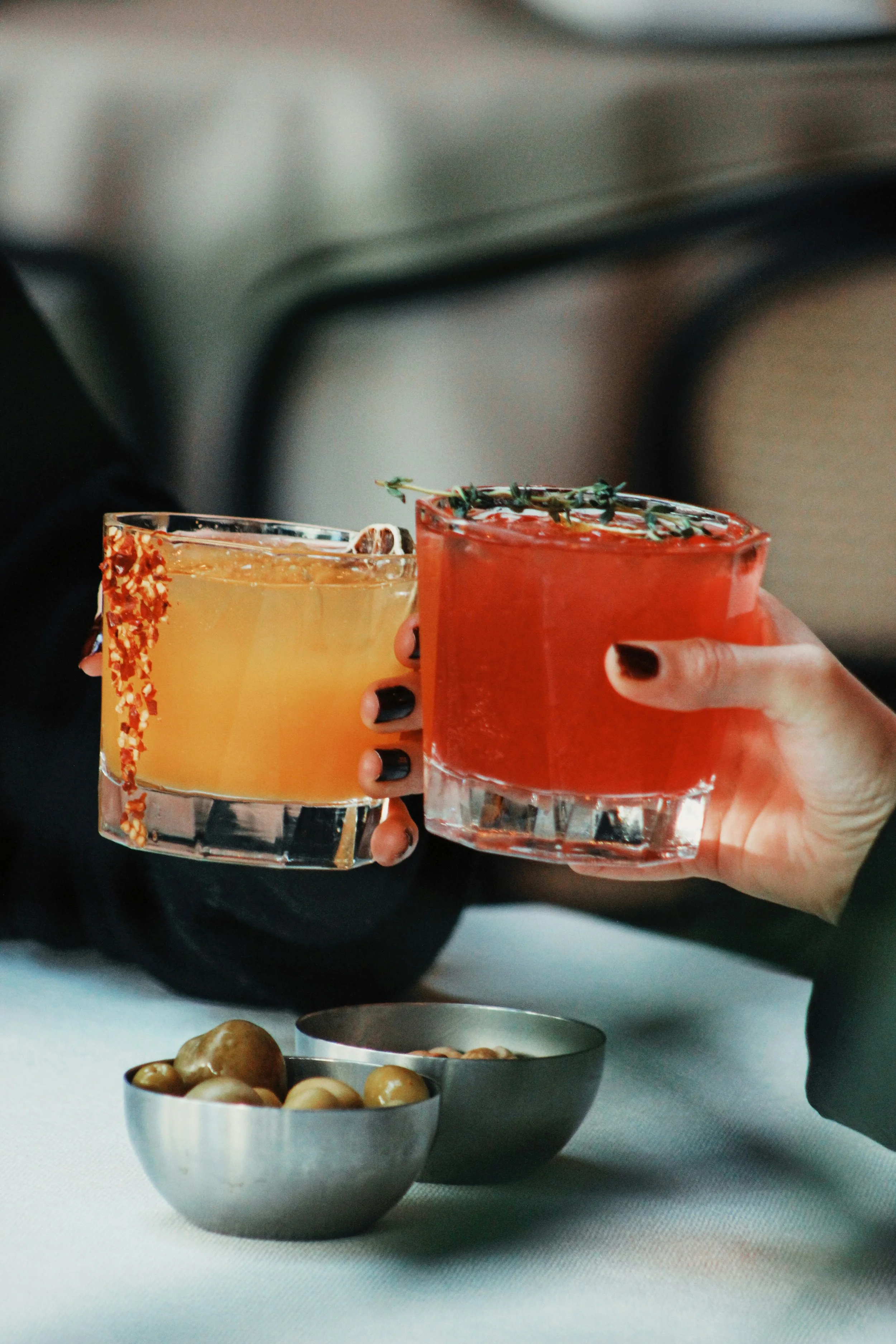 Two hands holding up glasses of cocktails, one yellow and one red, over a table with two small bowls of nuts.