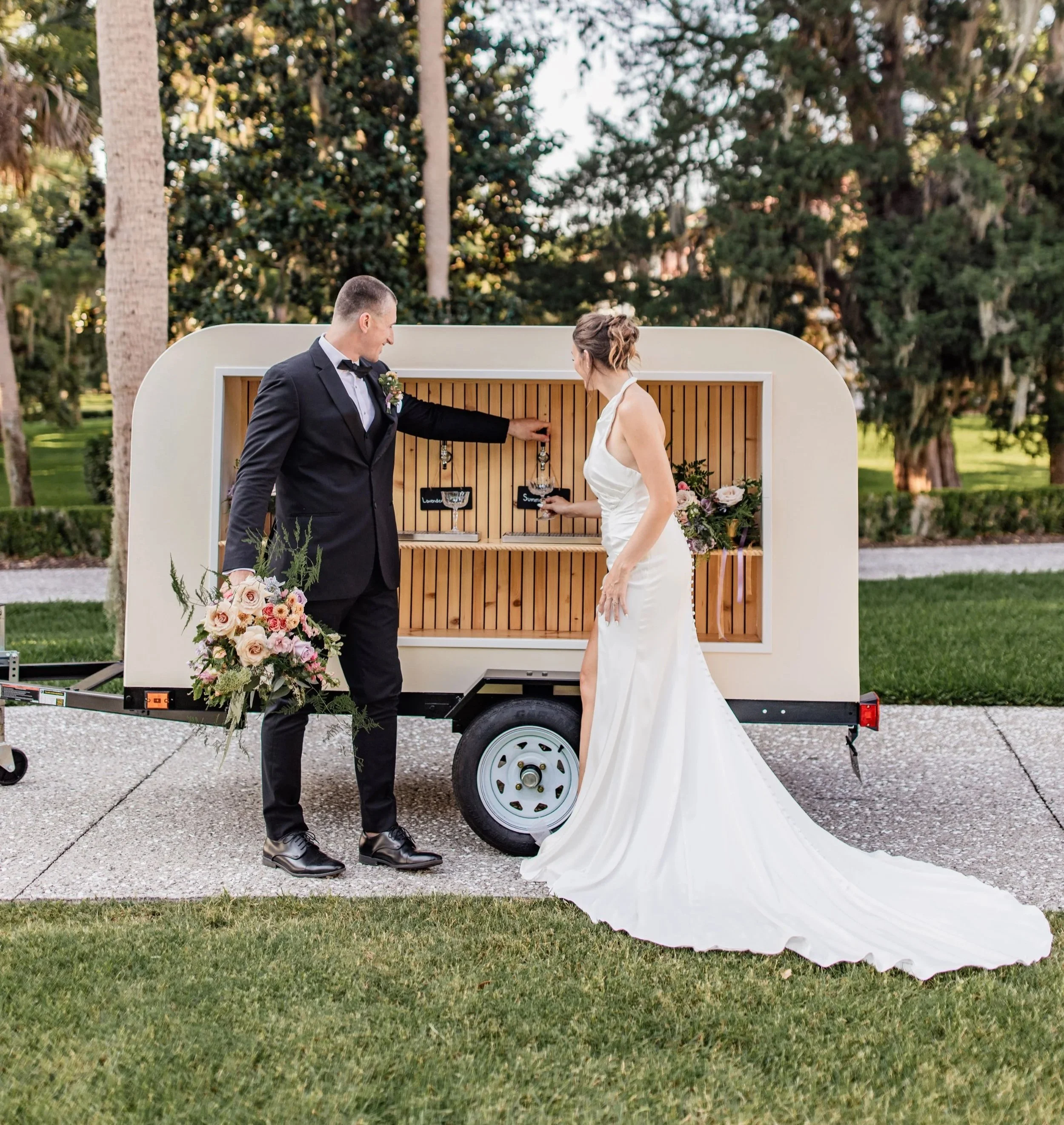 A bride and groom at their wedding reception, standing outside next to a small mobile bar with a wooden interior, the groom in a tuxedo holding a bouquet and the bride in a white wedding gown reaching for a drink.