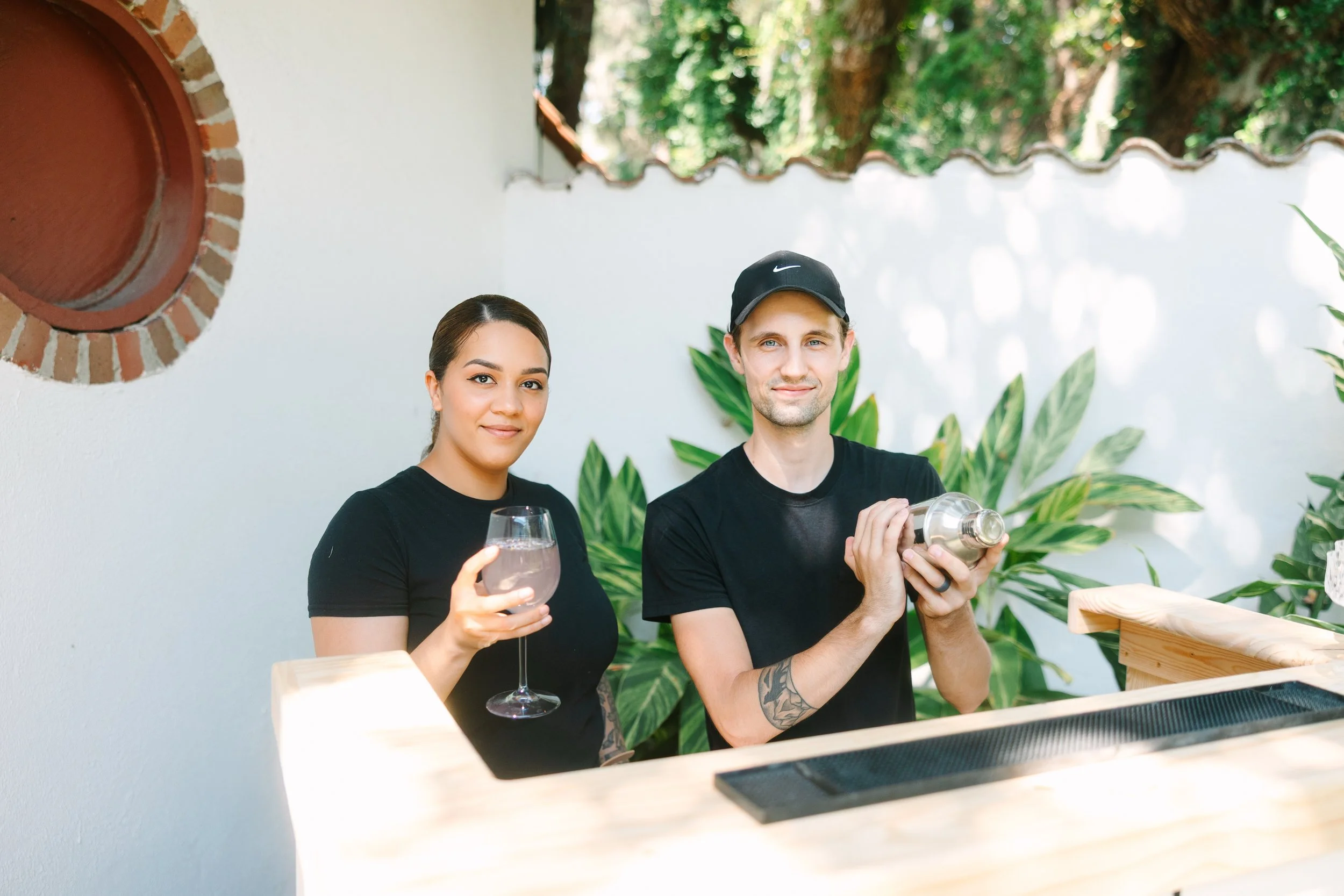 A woman holding a glass of clear drink and a man standing behind a bar with a cocktail shaker in hand. They are outdoors with green plants and a white wall in the background.