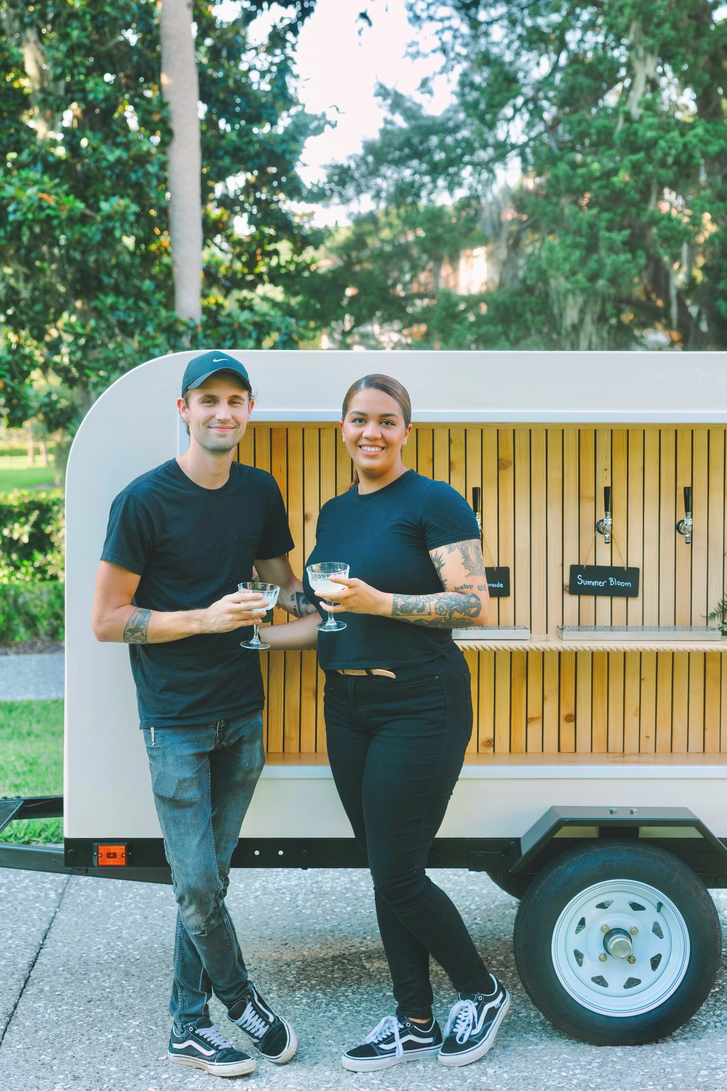 A young man and woman standing outdoors in front of a mobile beverage stand, holding glasses and smiling at the camera, with trees in the background.