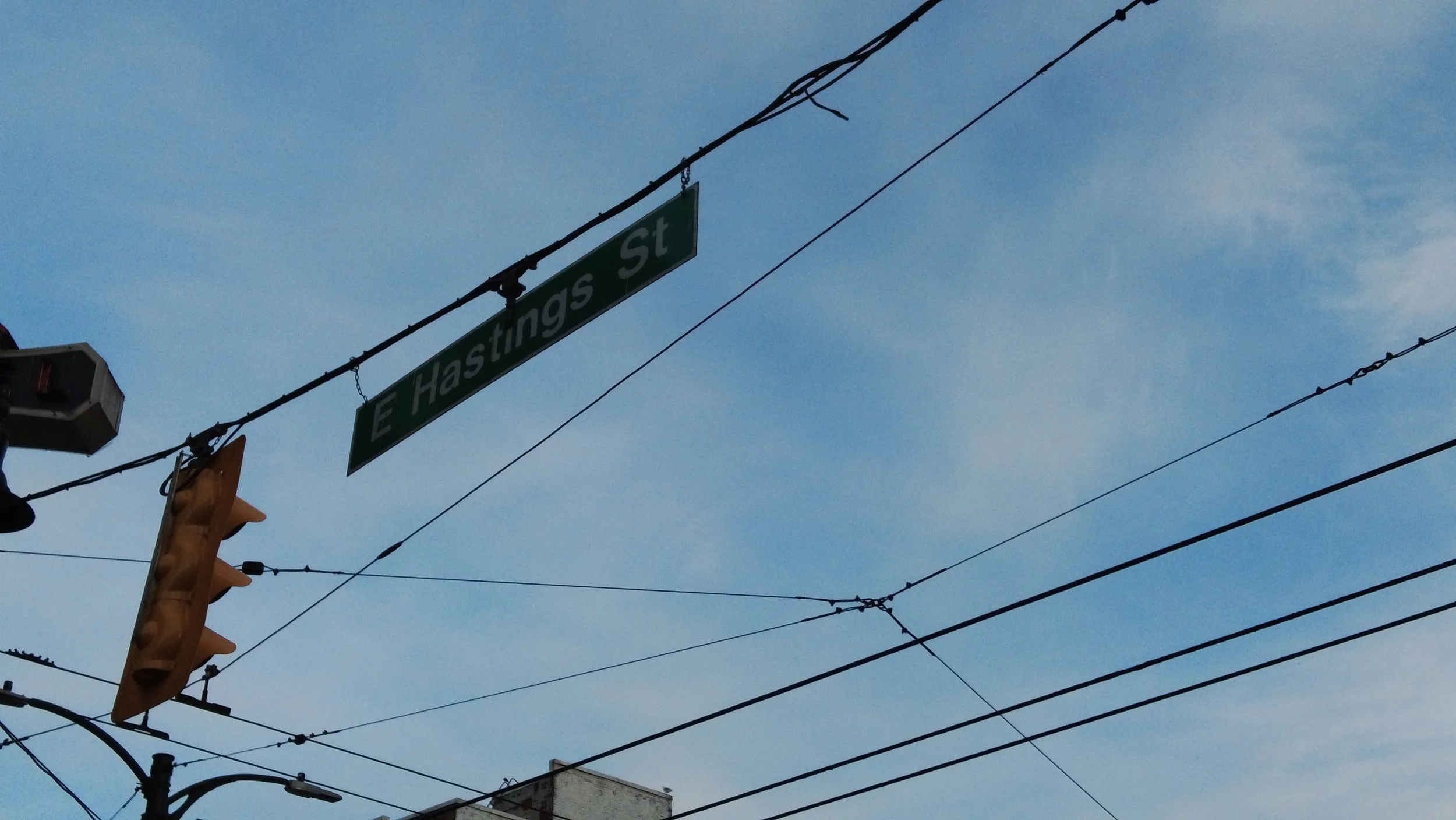 Street sign at E Hastings Street and a traffic light with overhead power lines against a blue sky.