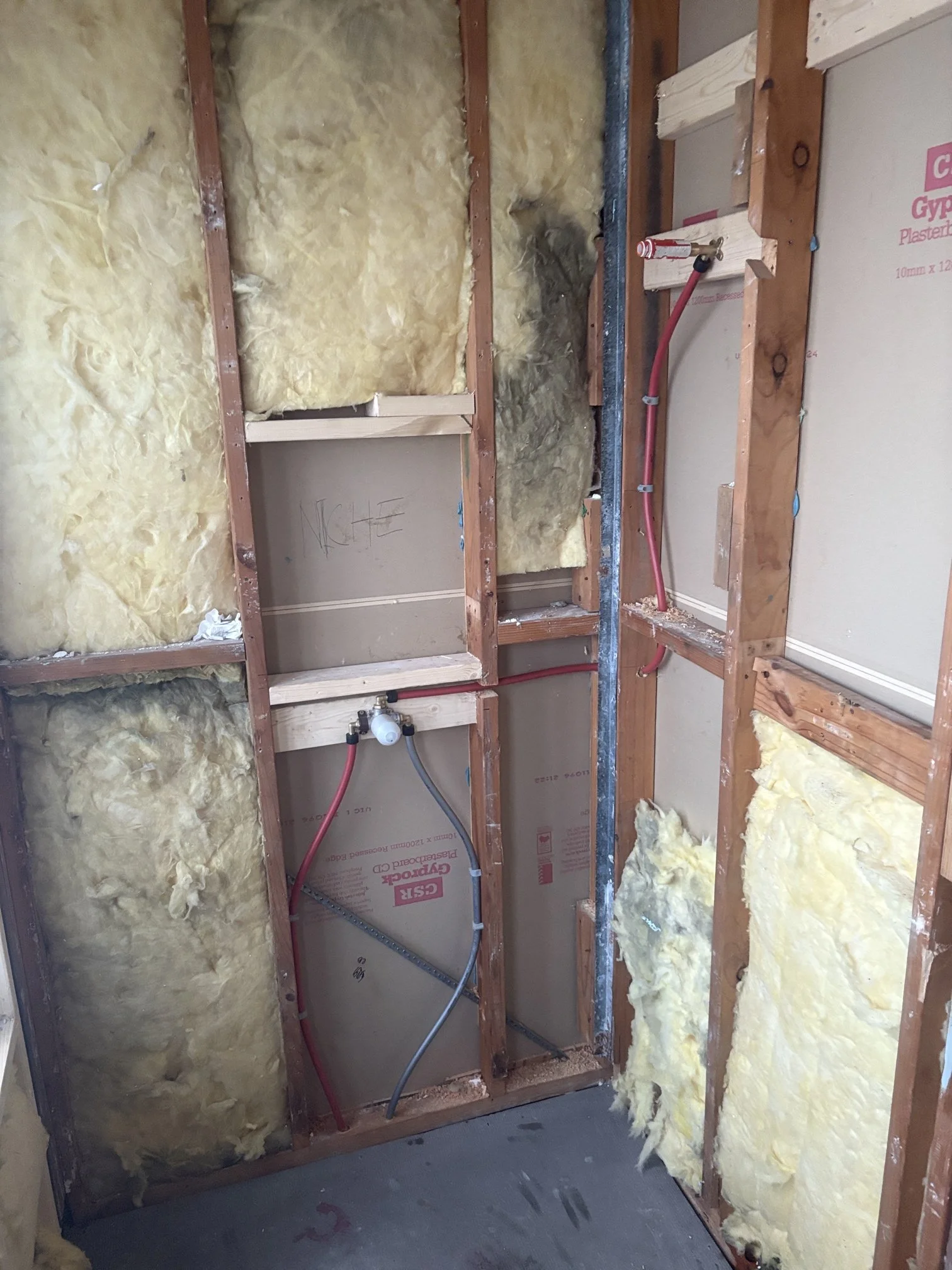 Part of a bathroom under construction showing insulation, exposed wooden framing, and plumbing pipes.