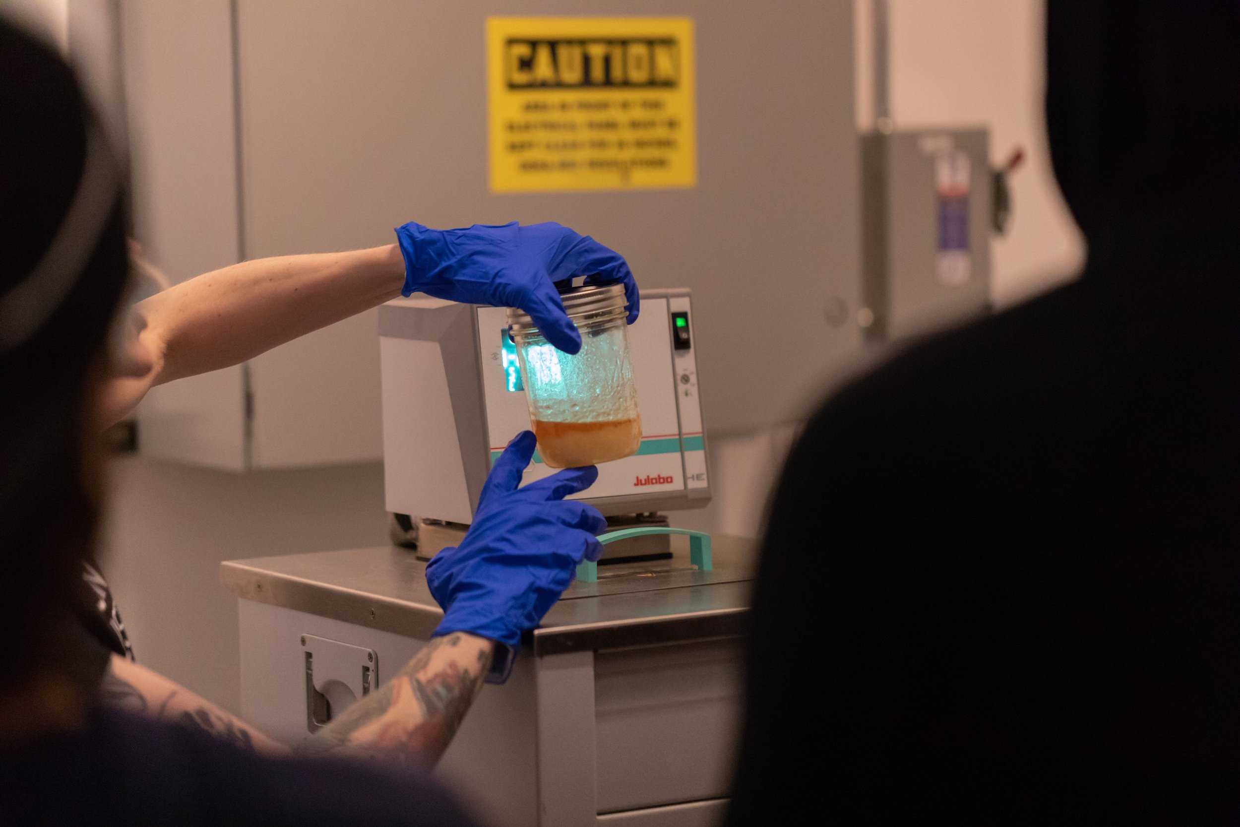 Person holding a jar with liquid in a laboratory setting, wearing blue gloves.