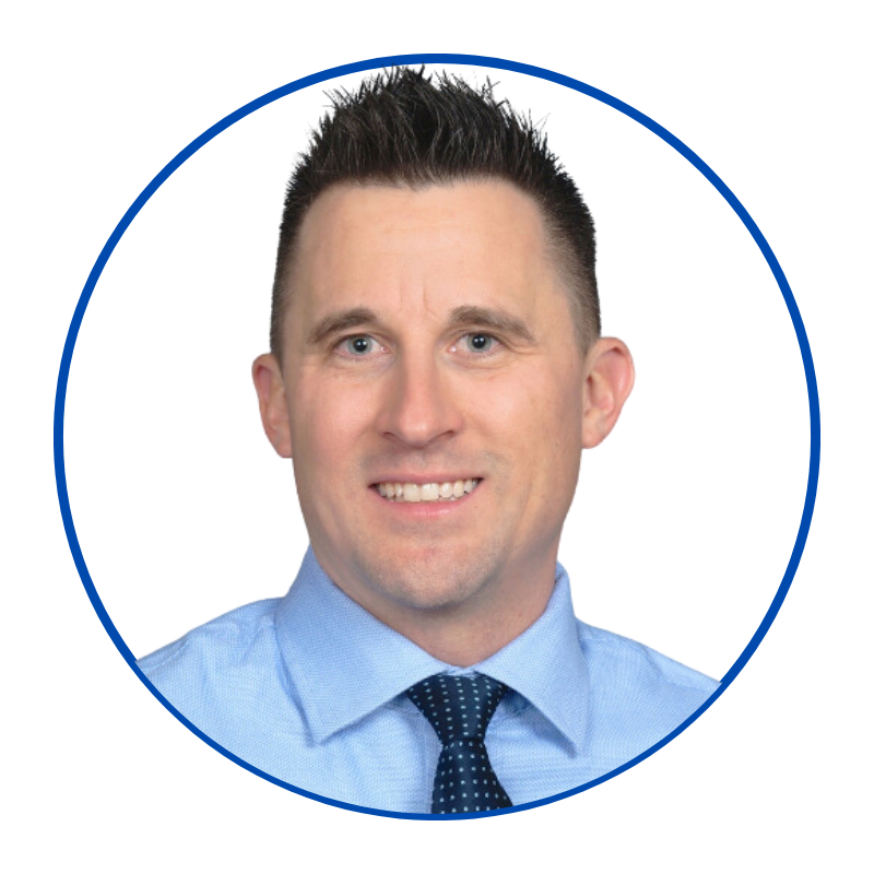 Professional headshot of a man with short dark hair, wearing a light blue collared shirt and a dark polka dot tie, set against a blue background.