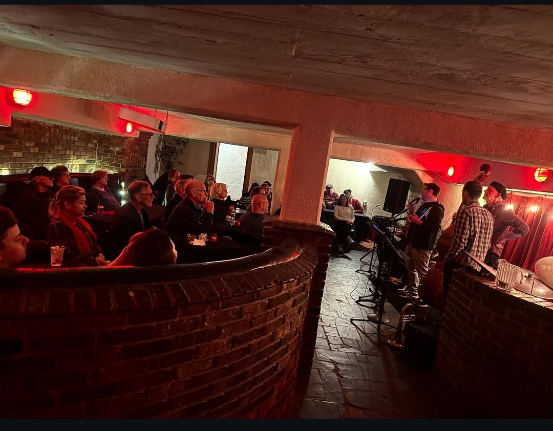 Underground room with brick walls, a projector screen displaying a video, purple ceiling lights, and chairs in the background.