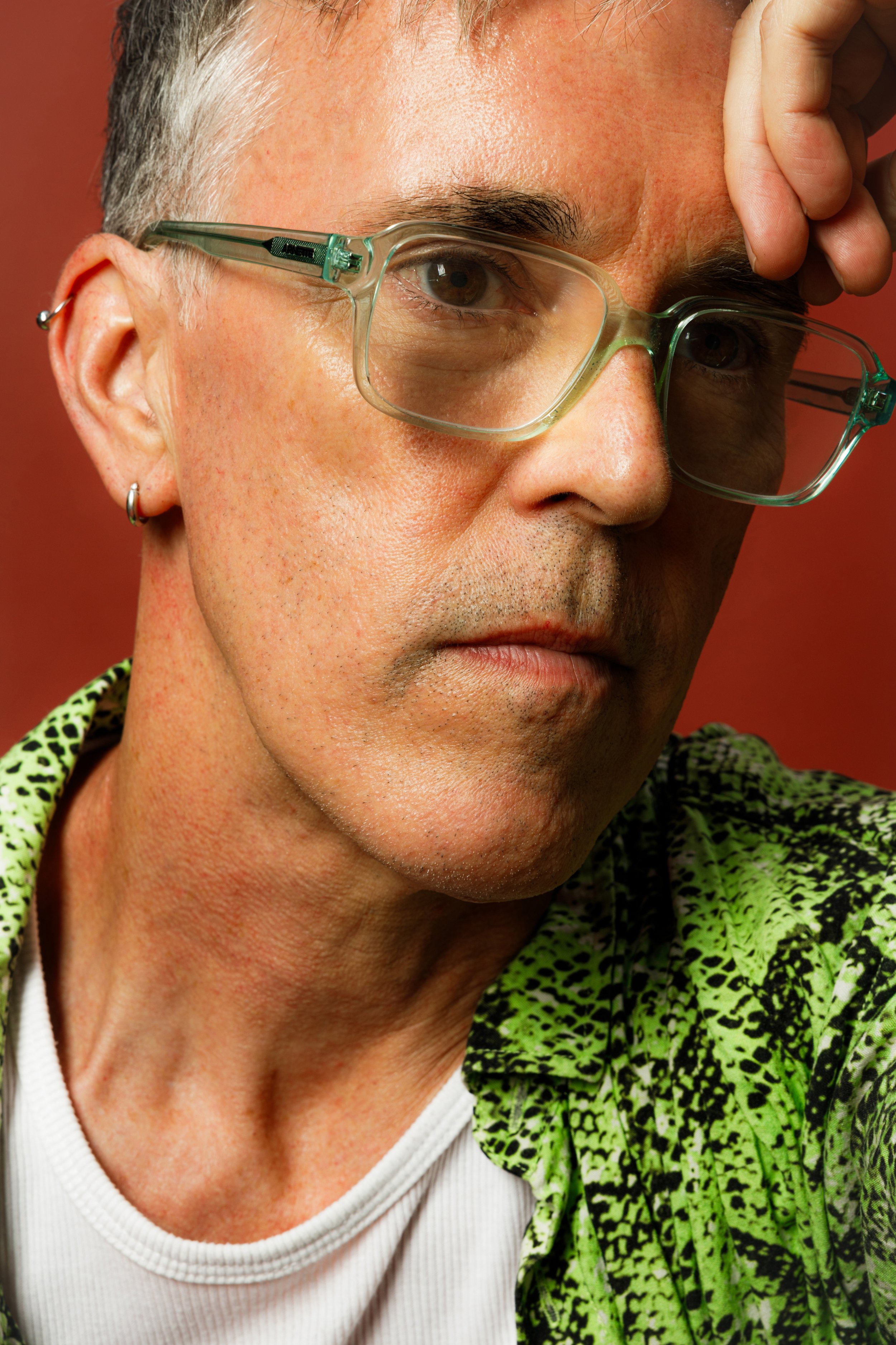 Close-up portrait of a person with short gray hair, wearing clear glasses, a small hoop earring, and a green and black patterned shirt. They are holding their forehead with one hand and looking directly at the camera.