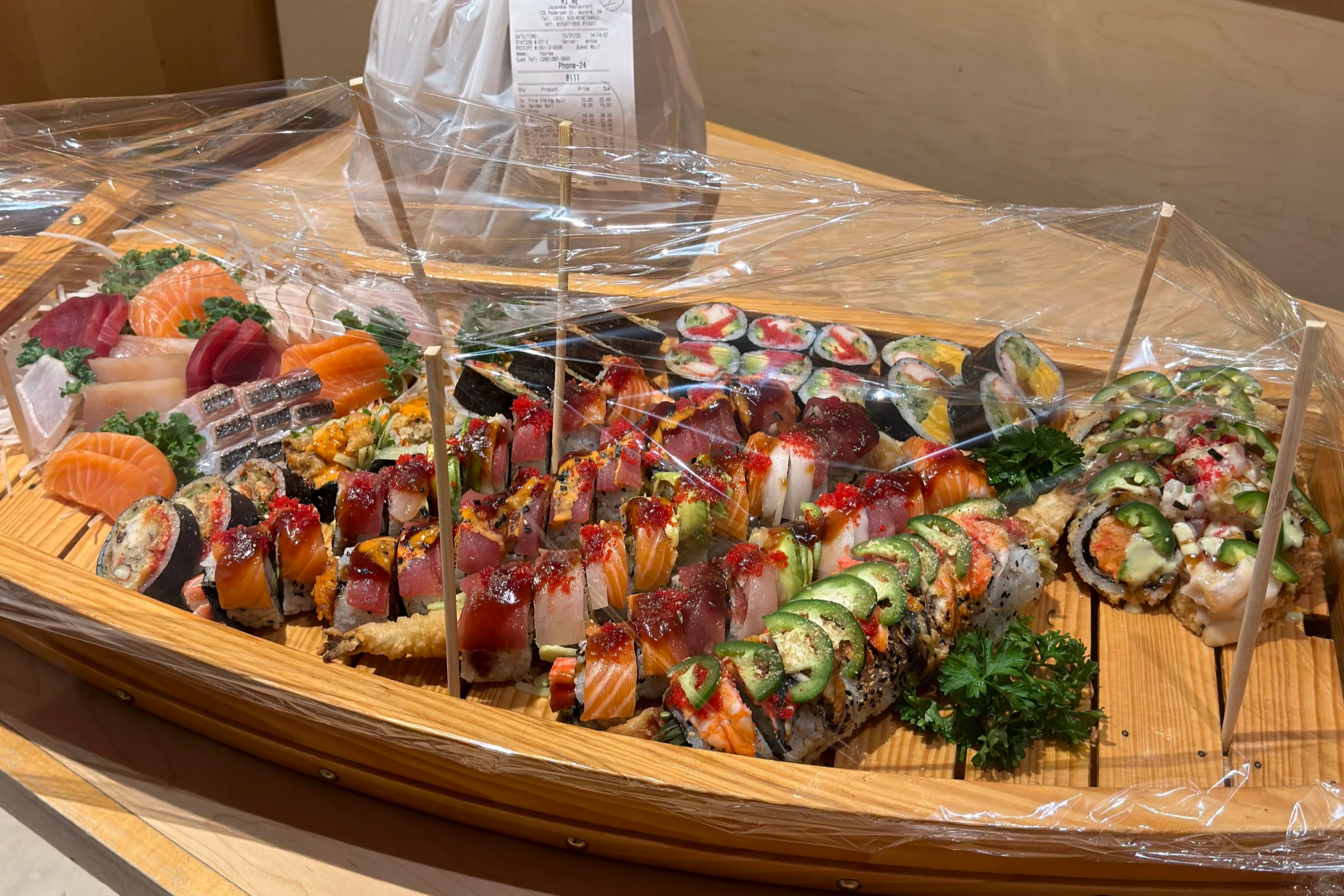 Assorted sushi and sashimi on a wooden boat with a clear plastic cover. Sushi boat, catering, events.