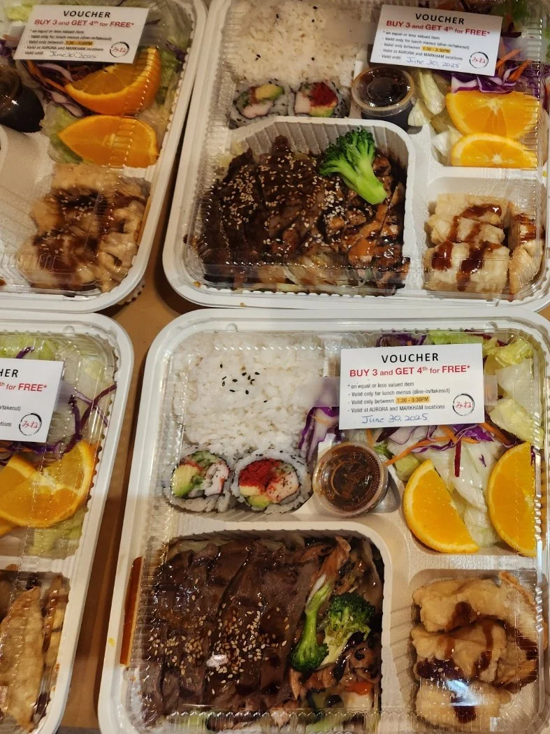 Multiple Japanese bento boxes containing sushi, rice, grilled meat with broccoli, and side salads with oranges, with vouchers on top. Bank brand lunch catering for employees.