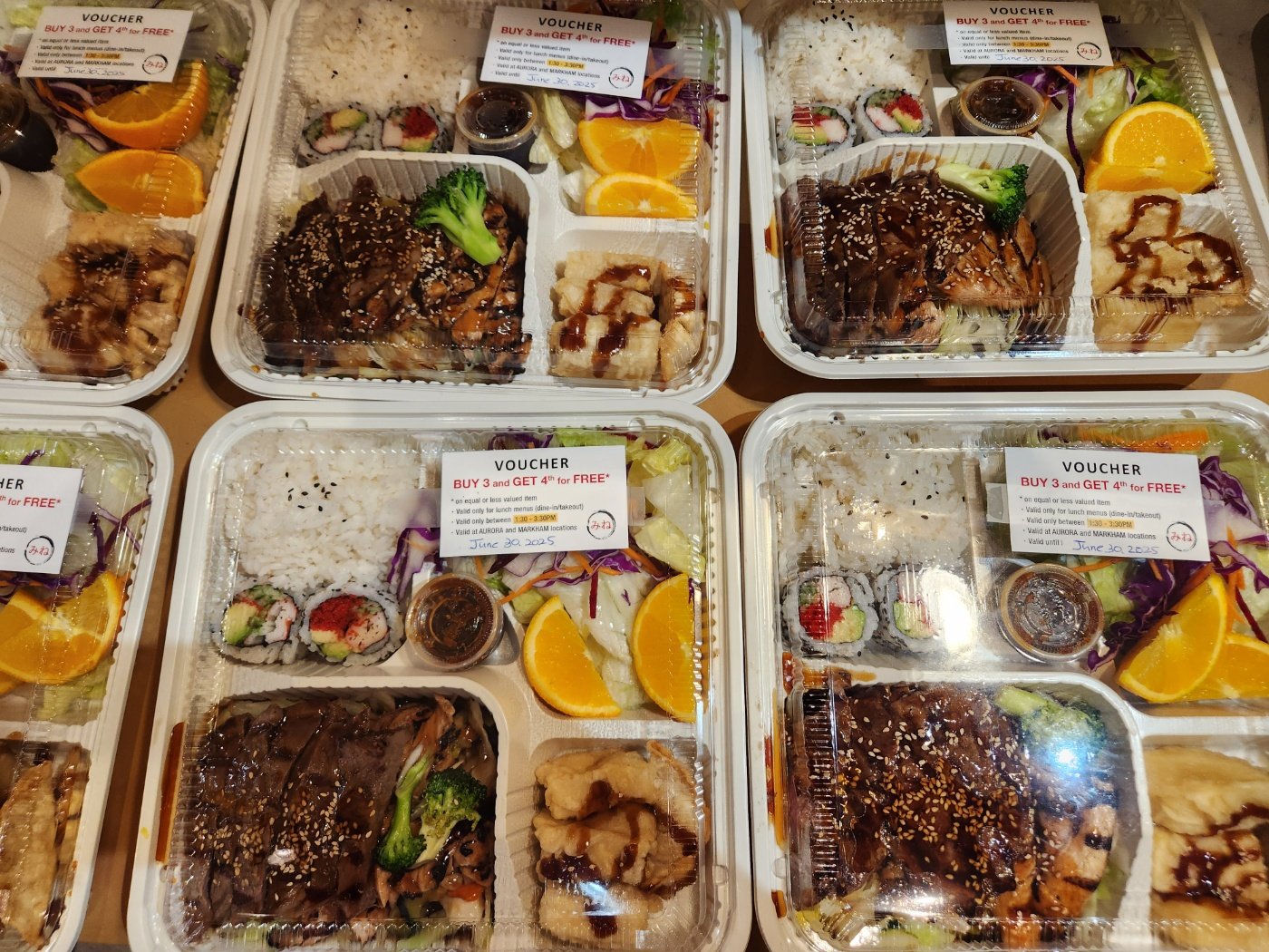 Multiple takeout sushi and Asian food meal trays with rice, sushi rolls, grilled chicken, broccoli, salad, orange slices, and sauce containers, labeled with vouchers for promotion.