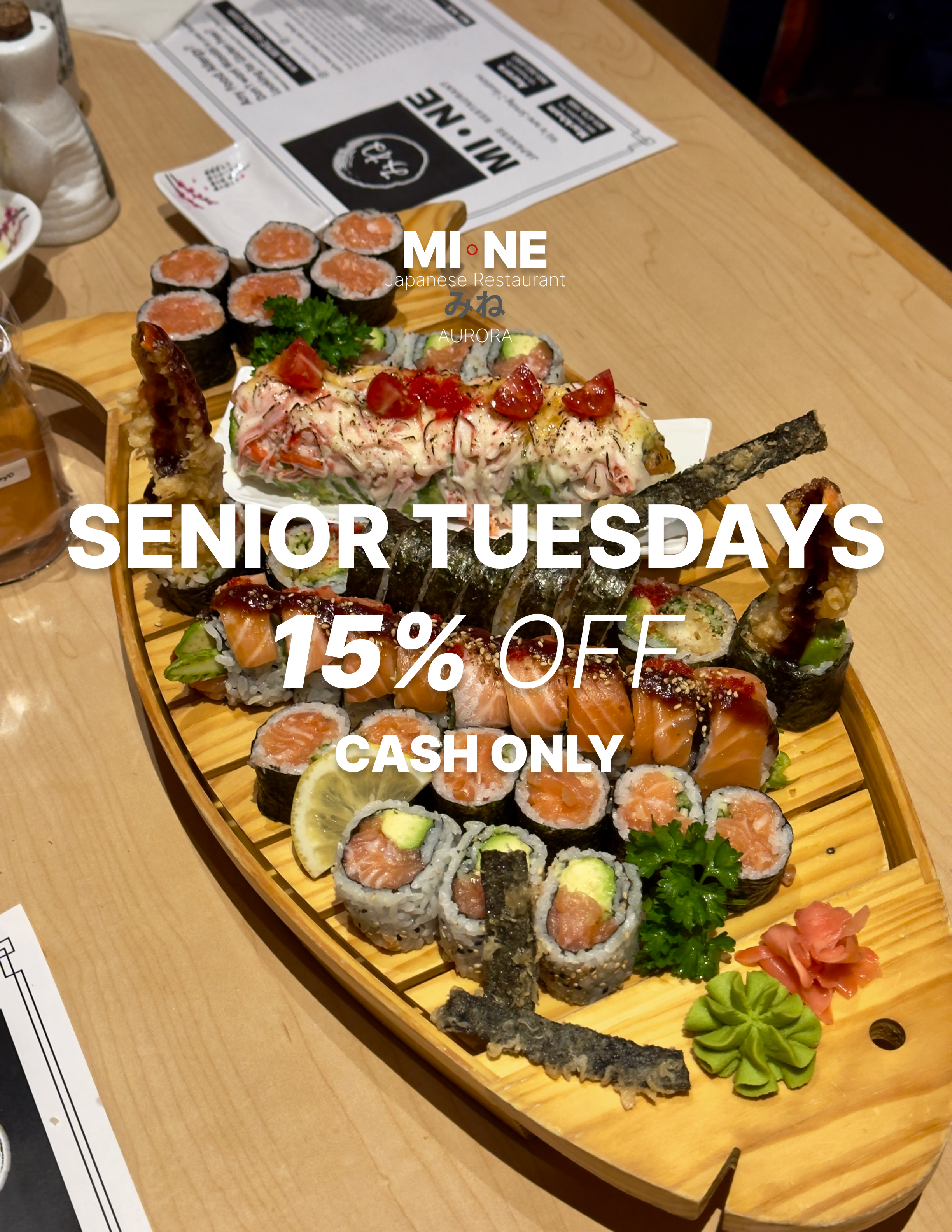 A wooden sushi platter with various types of sushi rolls, garnished with greens, wasabi, and pickled ginger, placed on a restaurant table. A menu is partially visible in the background.