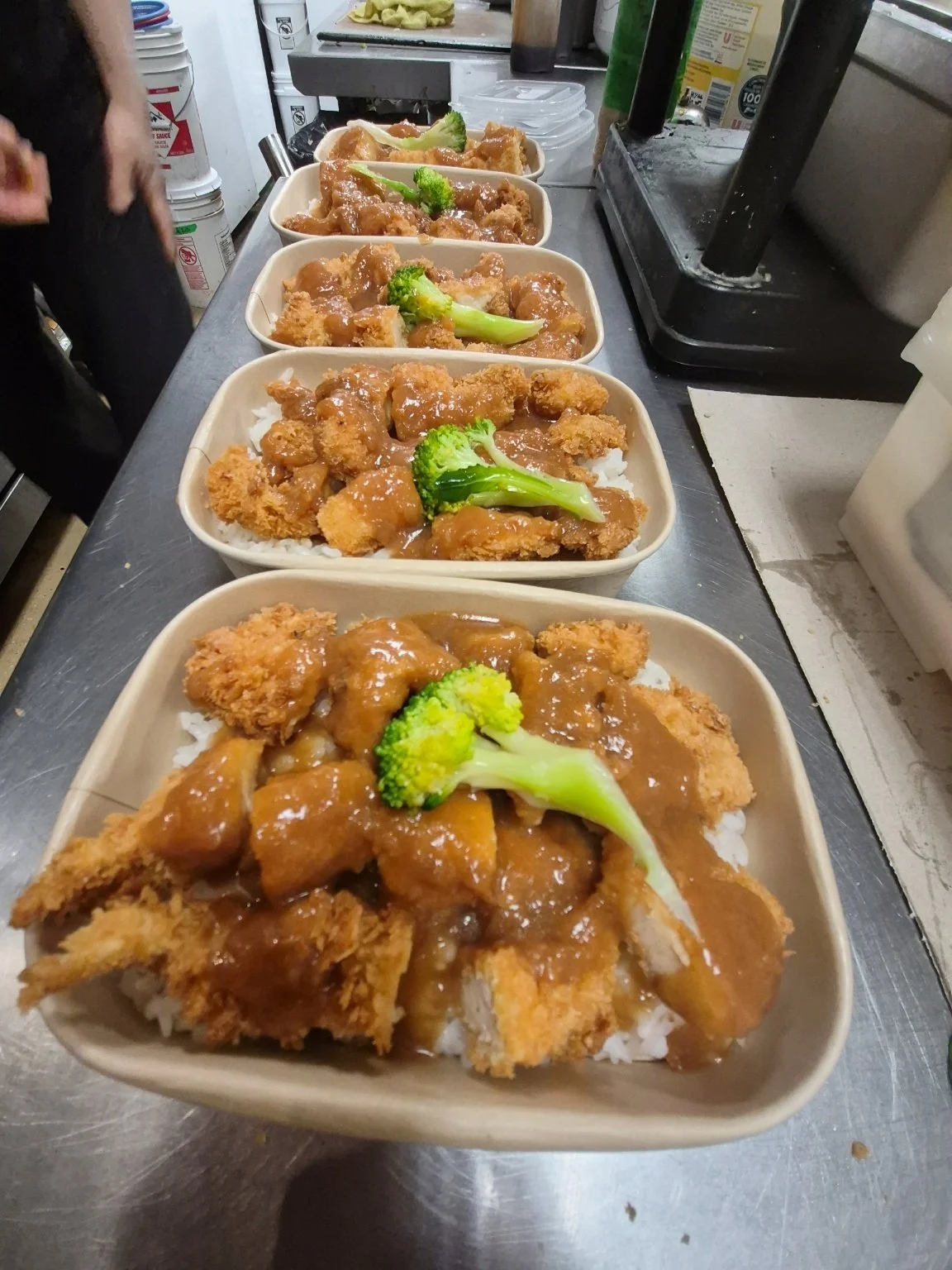Five takeout containers filled with fried chicken cutlets topped with brown gravy and broccoli served over white rice, arranged on a metal countertop in a kitchen.