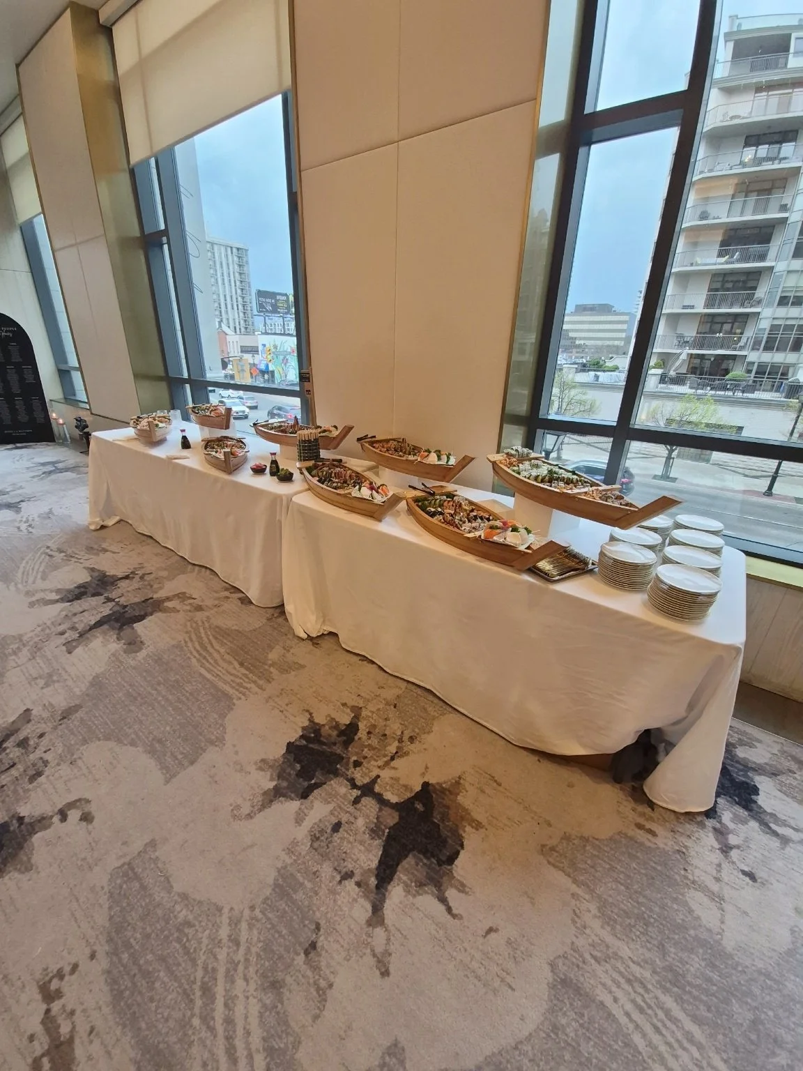 A buffet table set up in a modern hotel or conference center with a variety of sushi and other dishes displayed in boat-shaped trays. The table is covered with a white tablecloth, and there are stacks of plates near the right end of the table. Large 