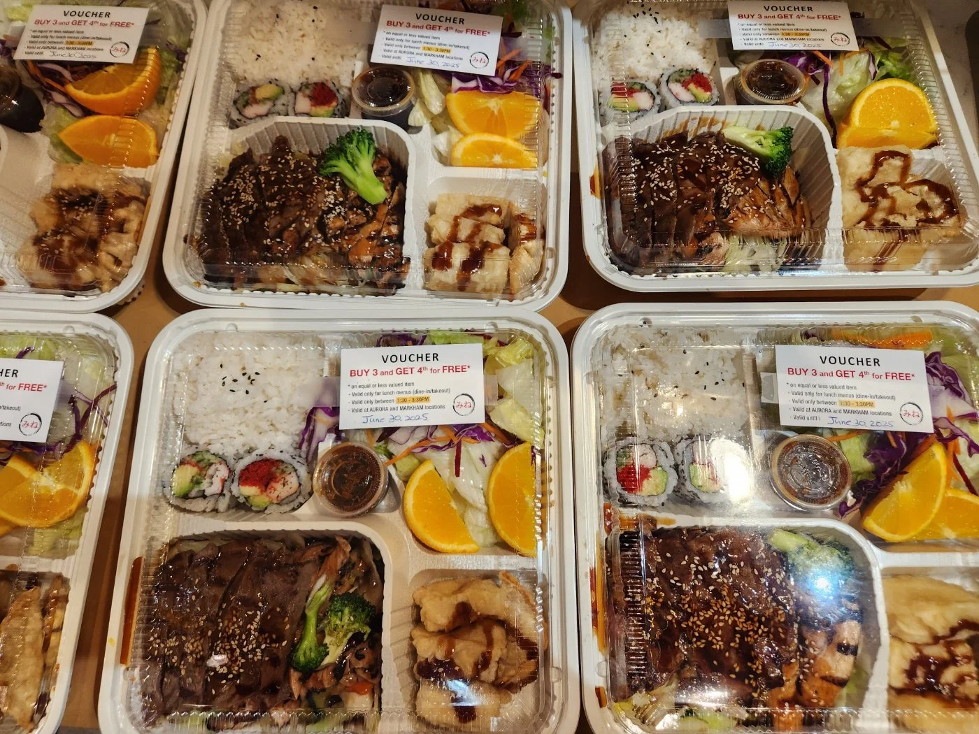 Pre-packaged Asian-style bento boxes with rice, sushi, grilled meat with sauce, mixed vegetables, orange slices, and dipping sauce.