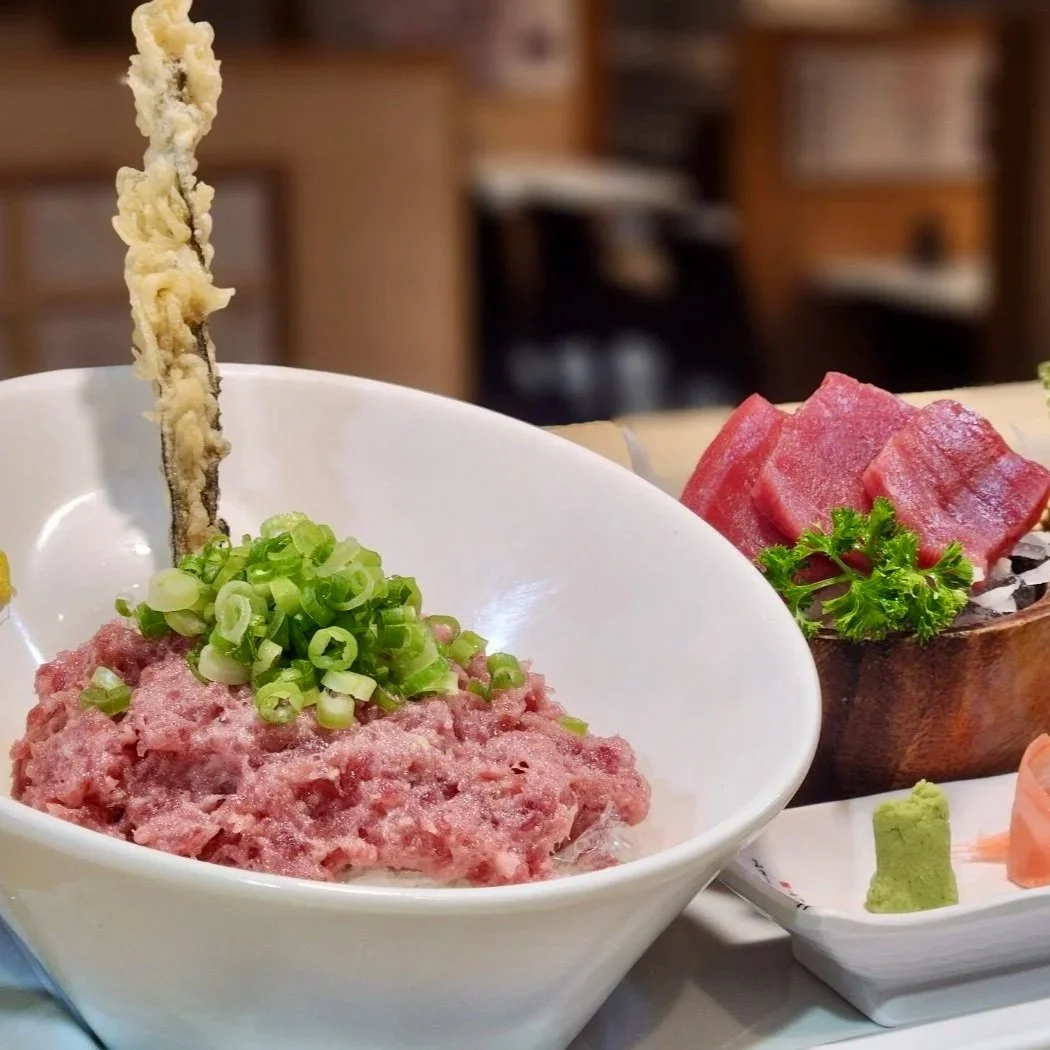 A bowl of chopped raw chu-toro bluefin tuna garnished with green onions. In the background, there are slices of raw bluefin tuna sashimi. There is also a small dish of wasabi and ginger.