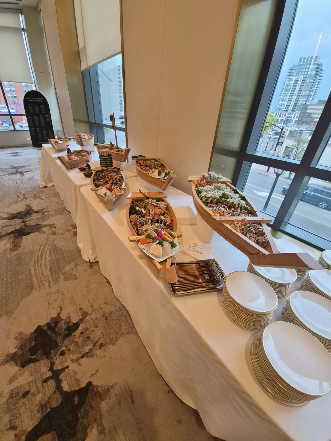 A buffet table set with various types of sushi and sashimi in small boats, with stacks of white plates nearby, in a room with large windows and cityscape views.