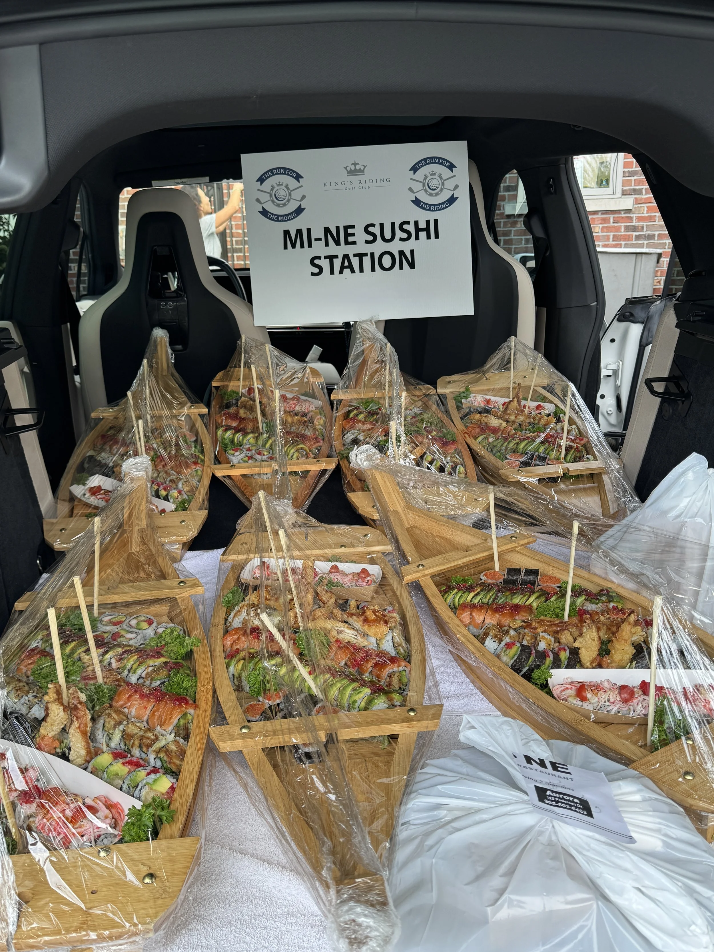 Multiple wooden boat-shaped sushi trays wrapped in plastic, displayed in the trunk of a vehicle for sushi station. Golf event catering sushi boats, takeout delivery.