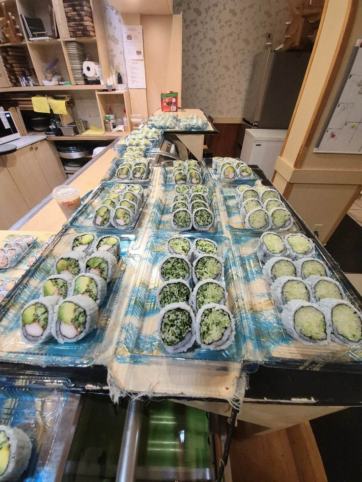 A variety of sushi rolls arranged on clear plastic trays on a counter in a kitchen, with a refrigerator and shelves in the background.