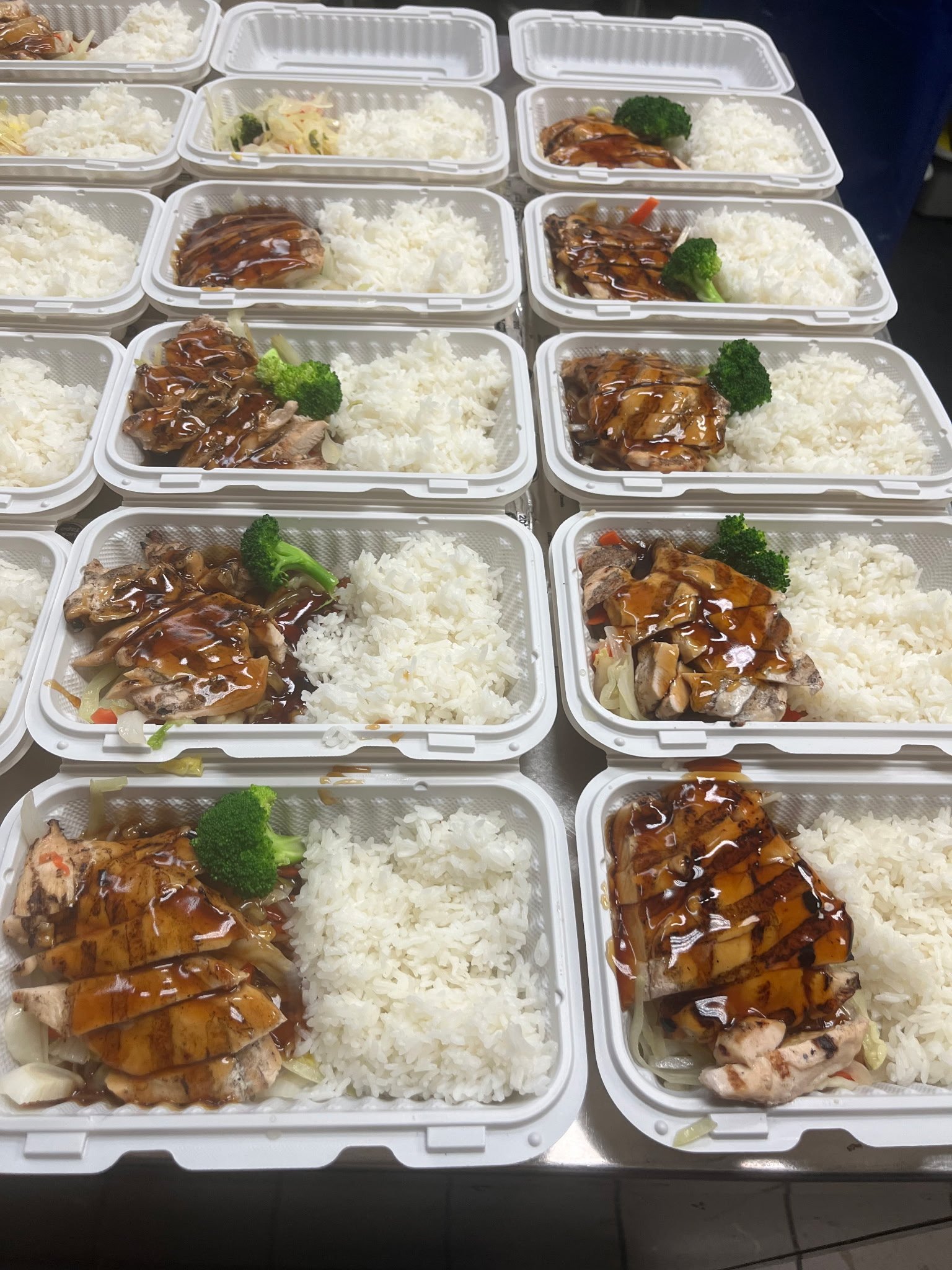 Multiple white meal containers filled with rice, grilled chicken with brown sauce, broccoli, and vegetable stir-fry, arranged on a table.