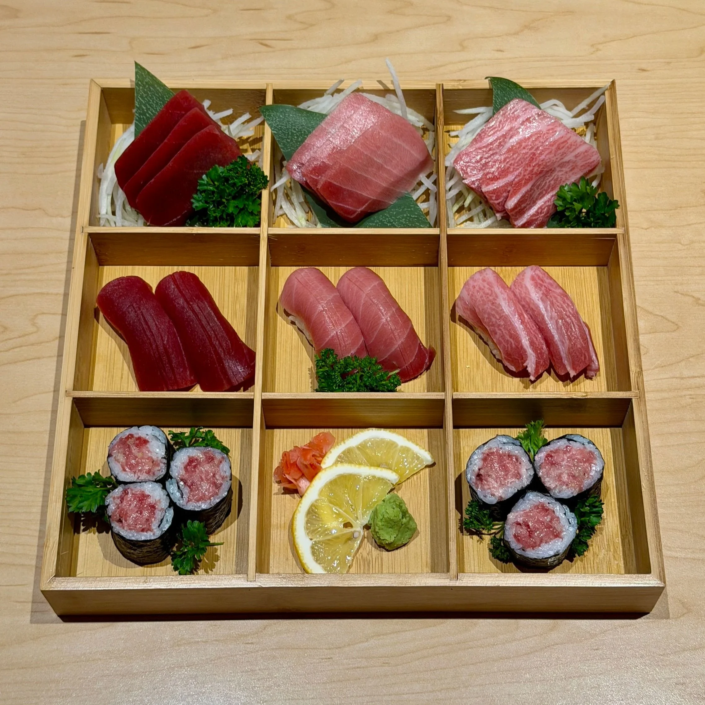 Assorted bluefin sashimi and sushi platter in a wooden box, featuring a fine selection of otoro, chu-toro and akami sashimi, nigiri, and sushi roll, garnished with lemon slices, wasabi and pickled ginger