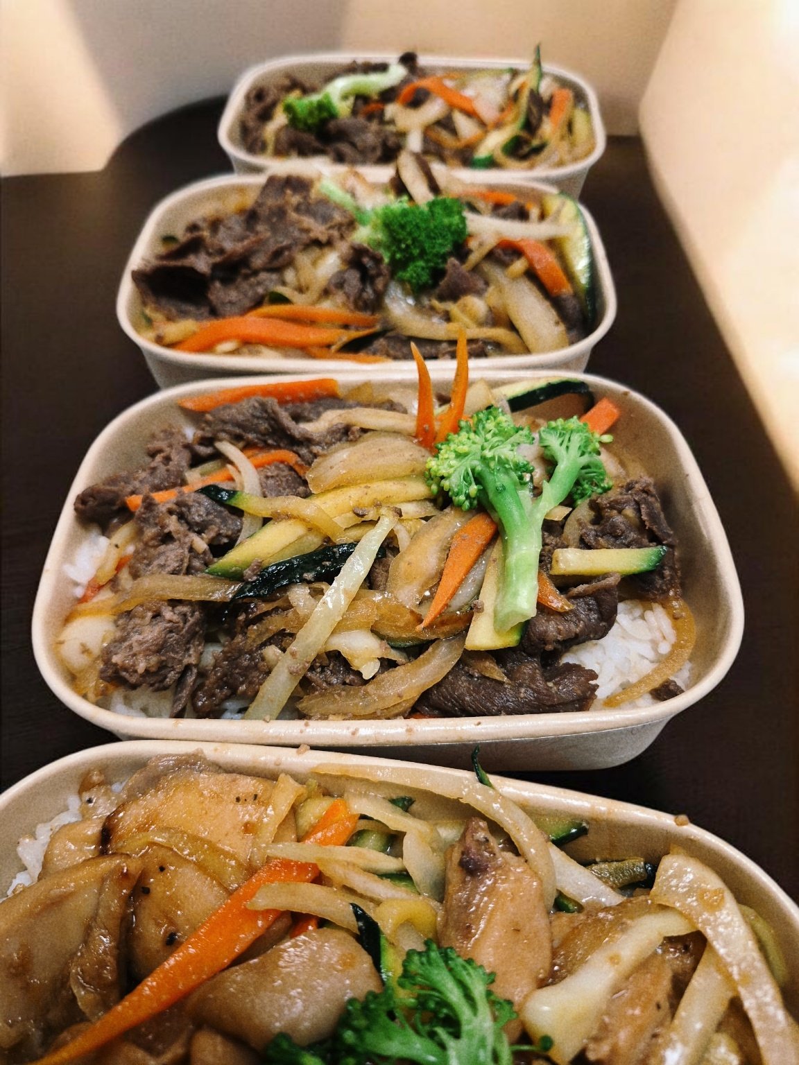 Bento boxes filled with rice, stir-fried vegetables, and sliced beef served in a row on a dark surface.