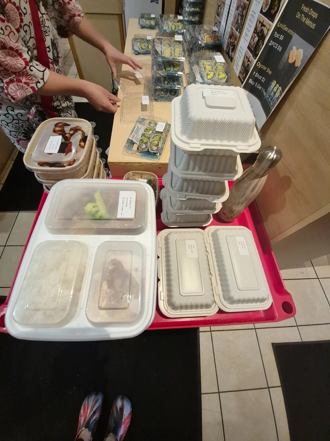 A sushi counter with multiple plastic containers of sushi, containers with food, a water bottle, and a menu board behind it. A person is reaching for some sushi.