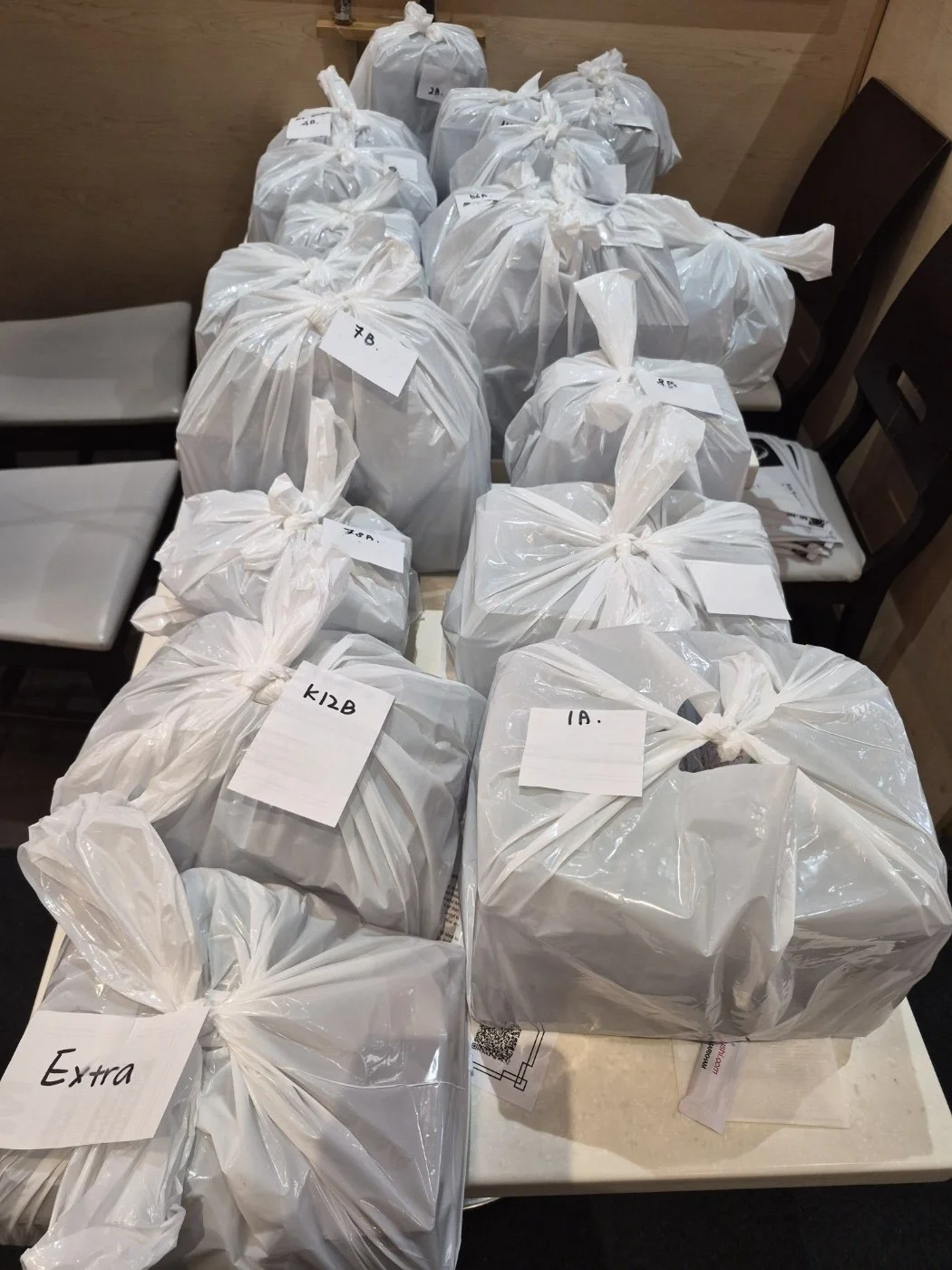 Multiple plastic bags filled with sushi and bento sets, labeled with handwritten tags. School lunch catering, takeout.