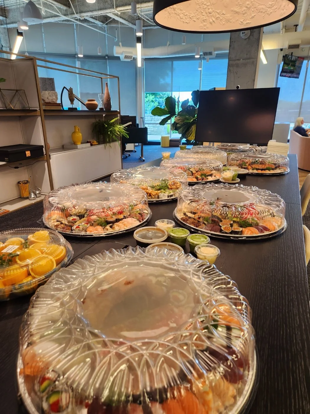 A long table set with multiple covered plastic trays of assorted sushi, small containers of wasabi and dipping sauces, and some orange and green desserts in a modern, brightly lit dining area.