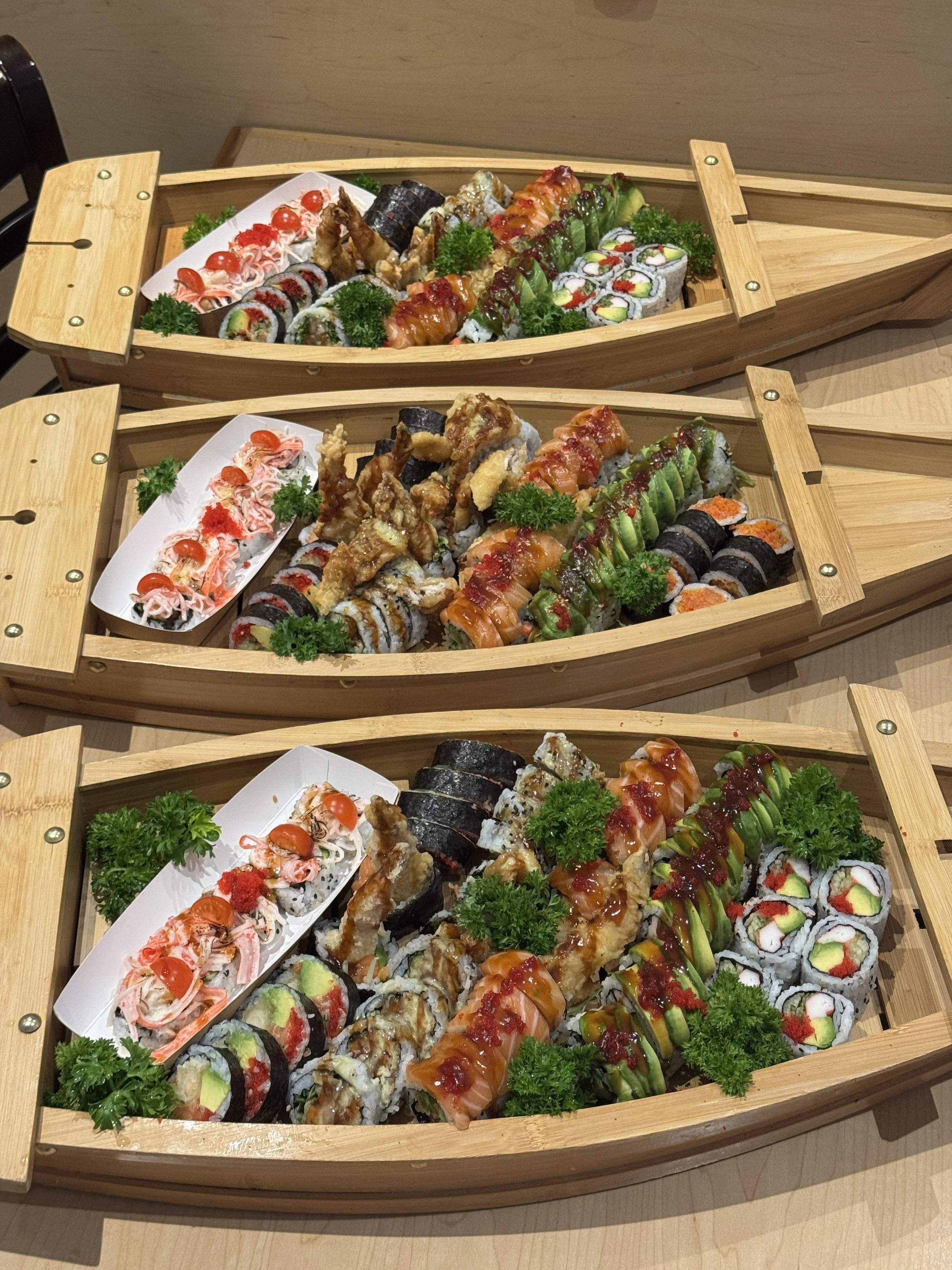 Three wooden boats filled with assorted colorful sushi rolls, garnished with parsley, on a wooden table. Sushi boat catering for corporate event, takeout.