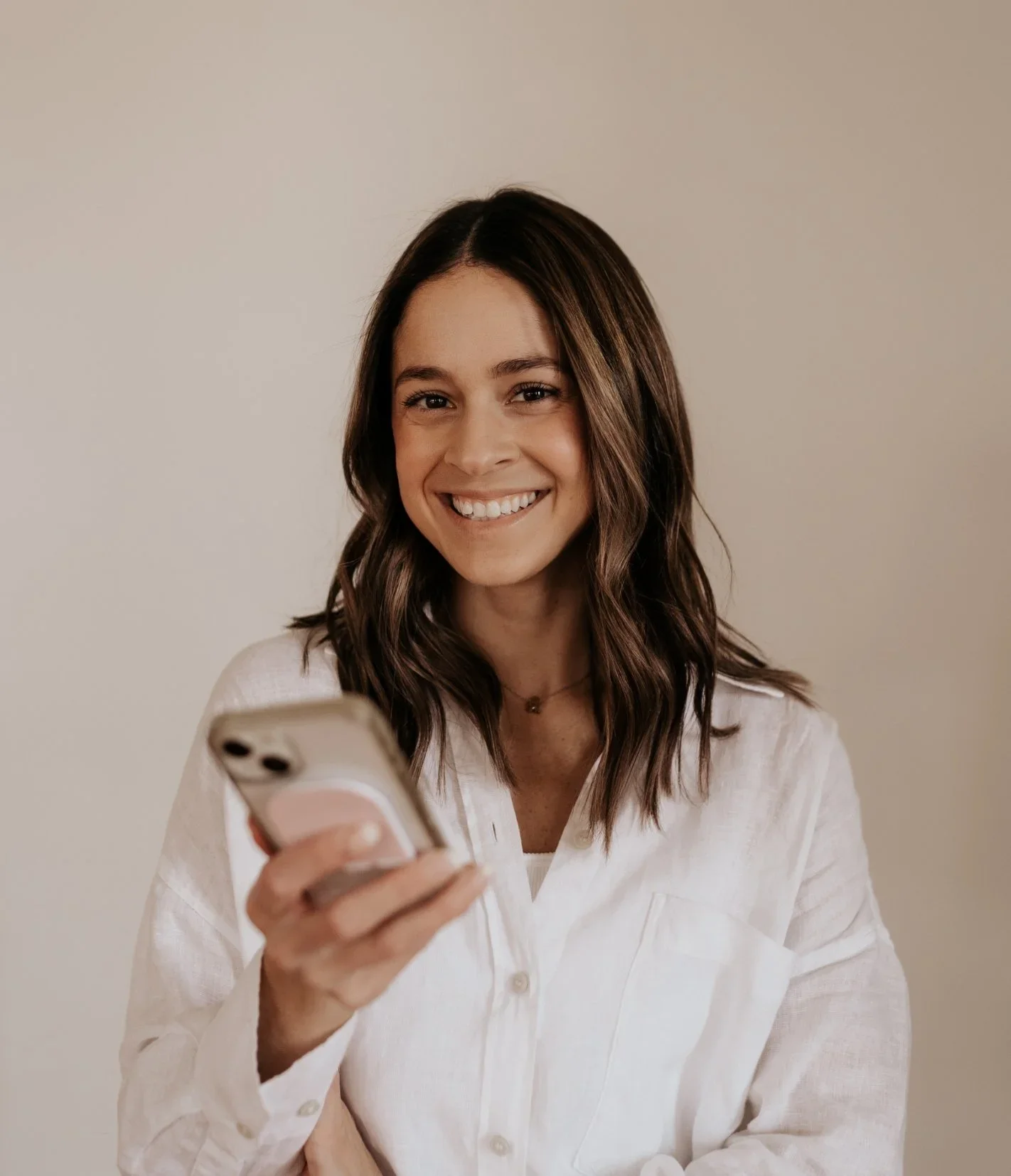 Kate Shewaga smiling and looking at her phone, wearing a white shirt and standing against a plain background.