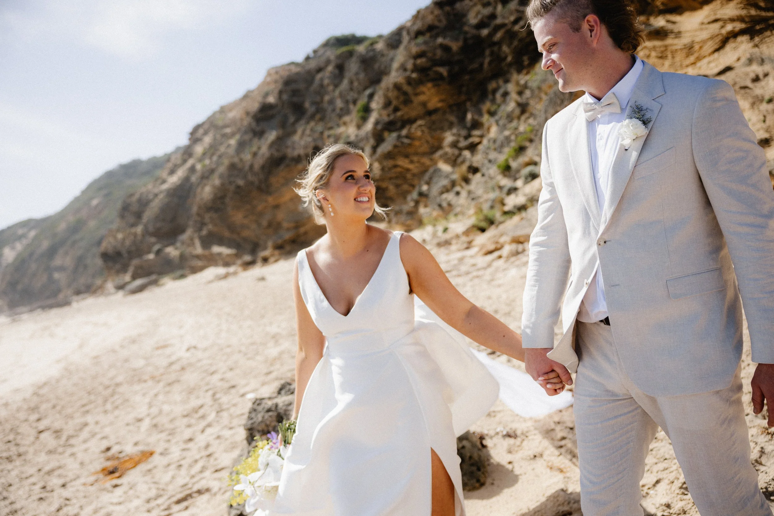 A newlywed couple kissing on the beach, with the bride holding a bouquet of flowers, wearing a white wedding gown, and the groom in a light-colored suit, with the ocean and rocks in the background.