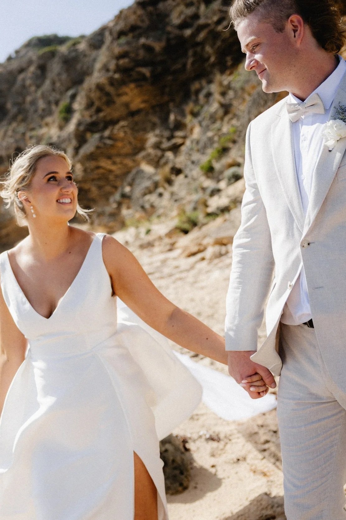 A newlywed couple kissing on the beach, with the bride holding a bouquet of flowers, wearing a white wedding gown, and the groom in a light-colored suit, with the ocean and rocks in the background.