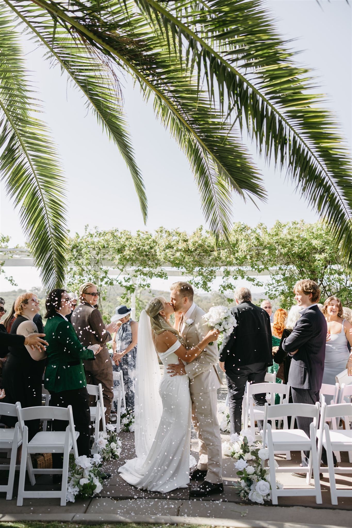 Couple in wedding attire sharing a kiss outdoors, with the bride in a white dress and long veil, and the groom in a light-colored suit, surrounded by greenery.
