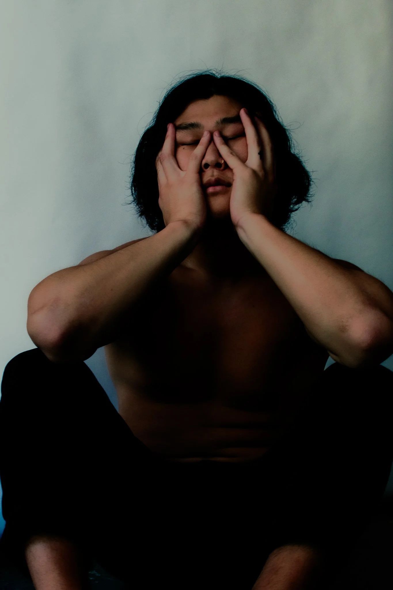Person with long dark hair and tan skin sitting shirtless against a plain wall, touching their face with both hands, eyes closed.