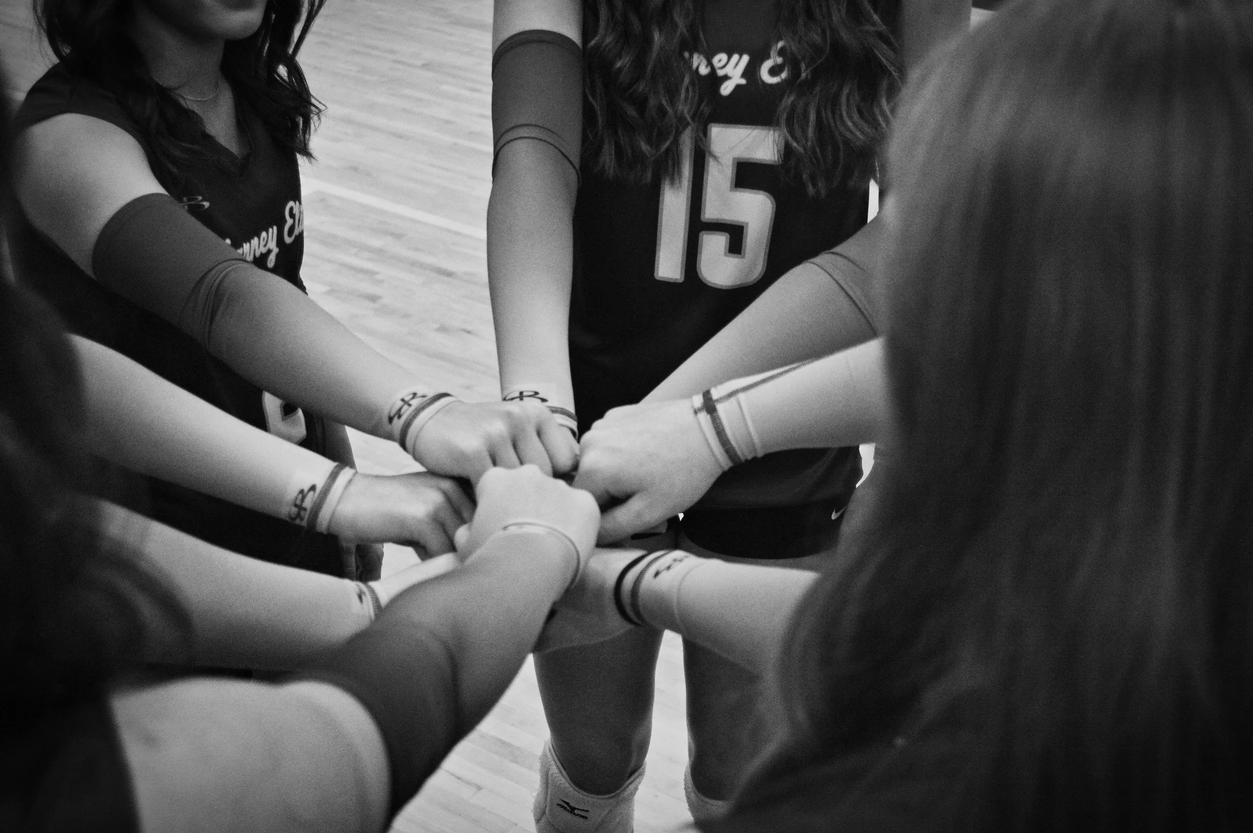 A group of female volleyball players in black jerseys with their arms stacked together in a huddle.