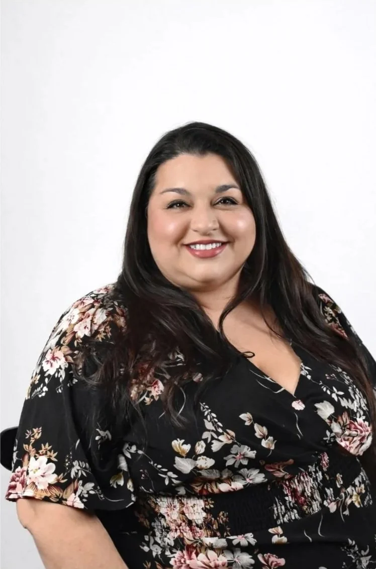 A woman with long black hair and red lipstick smiling in front of a colorful graffiti wall, wearing a black top with a sheer polka dot neckline.
