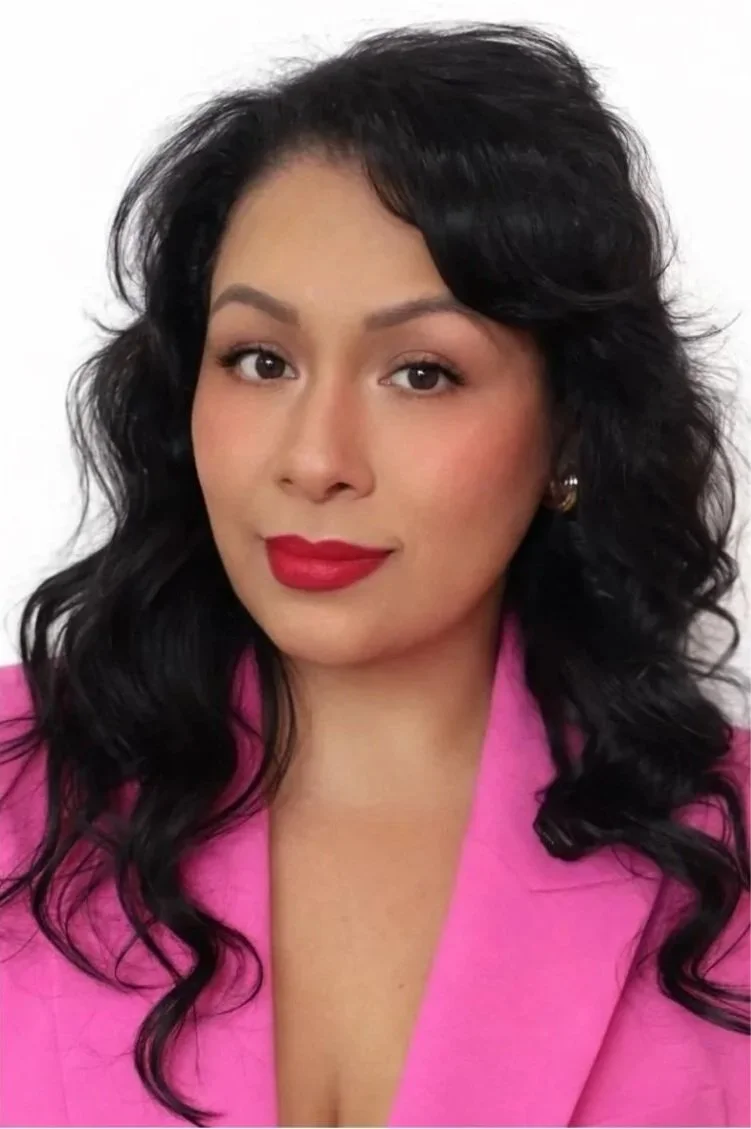A woman with black, wavy hair and a bright smile wearing a red satin dress with a one-shoulder design and sparkling jewelry, including earrings and a necklace, posing indoors with a blurred modern background.